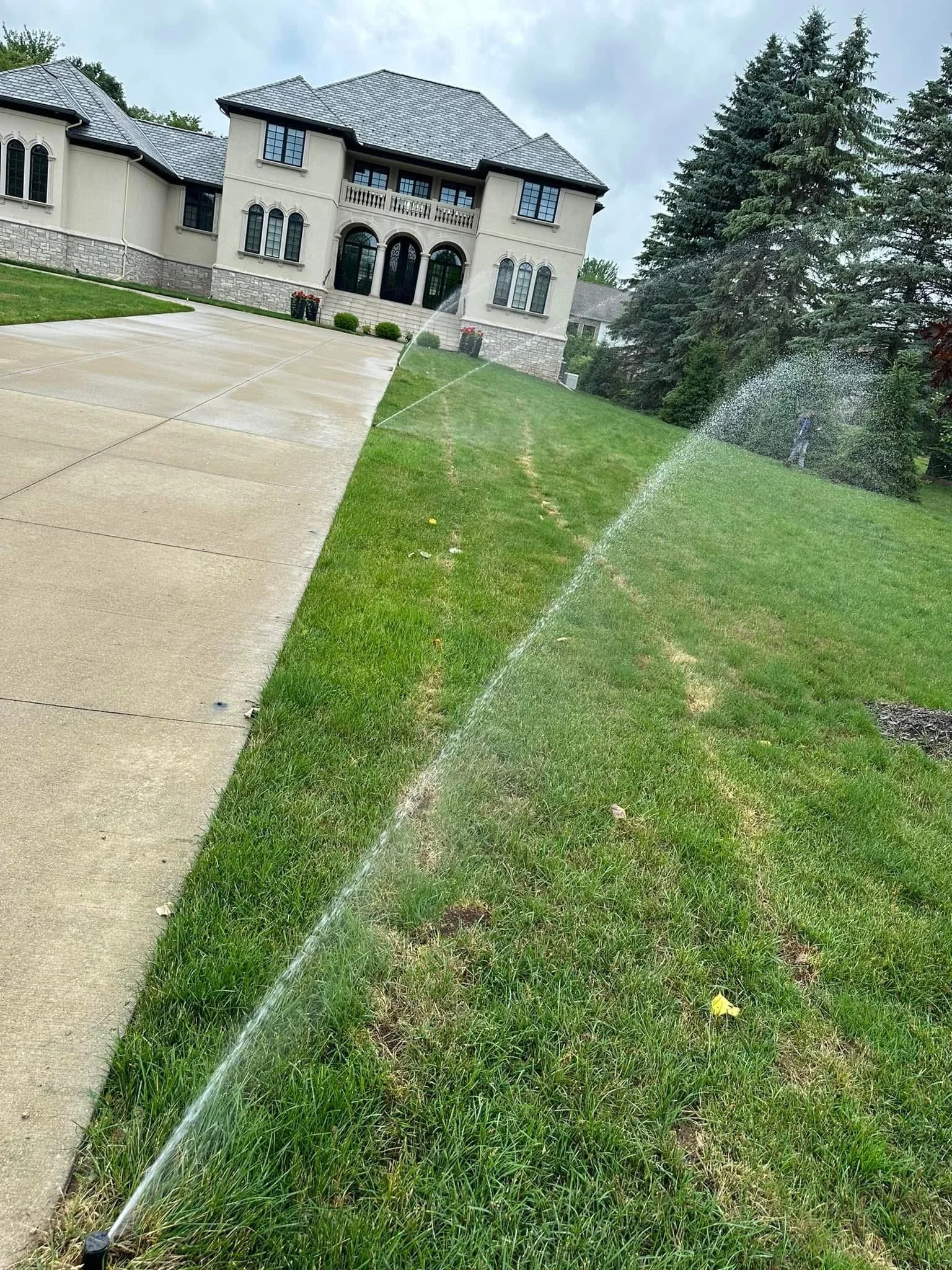 A house with a gray tiled roof and beige exterior walls, featuring arched windows and a balcony, is seen behind a green lawn and a concrete driveway. Sprinklers are watering the grass on a cloudy day.