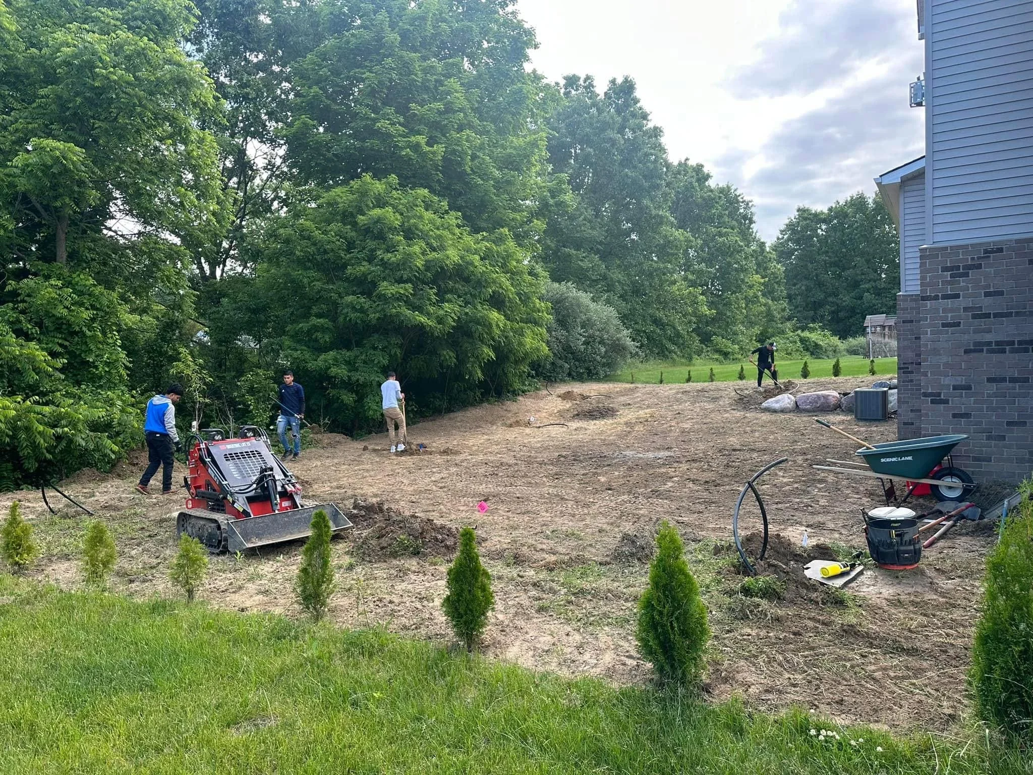 Several people are working on landscaping a yard near a blue house with brick foundation. There are trees and bushes on the left and a grassy area with small shrubs on the right. The yard has dirt and construction tools scattered around, including a 