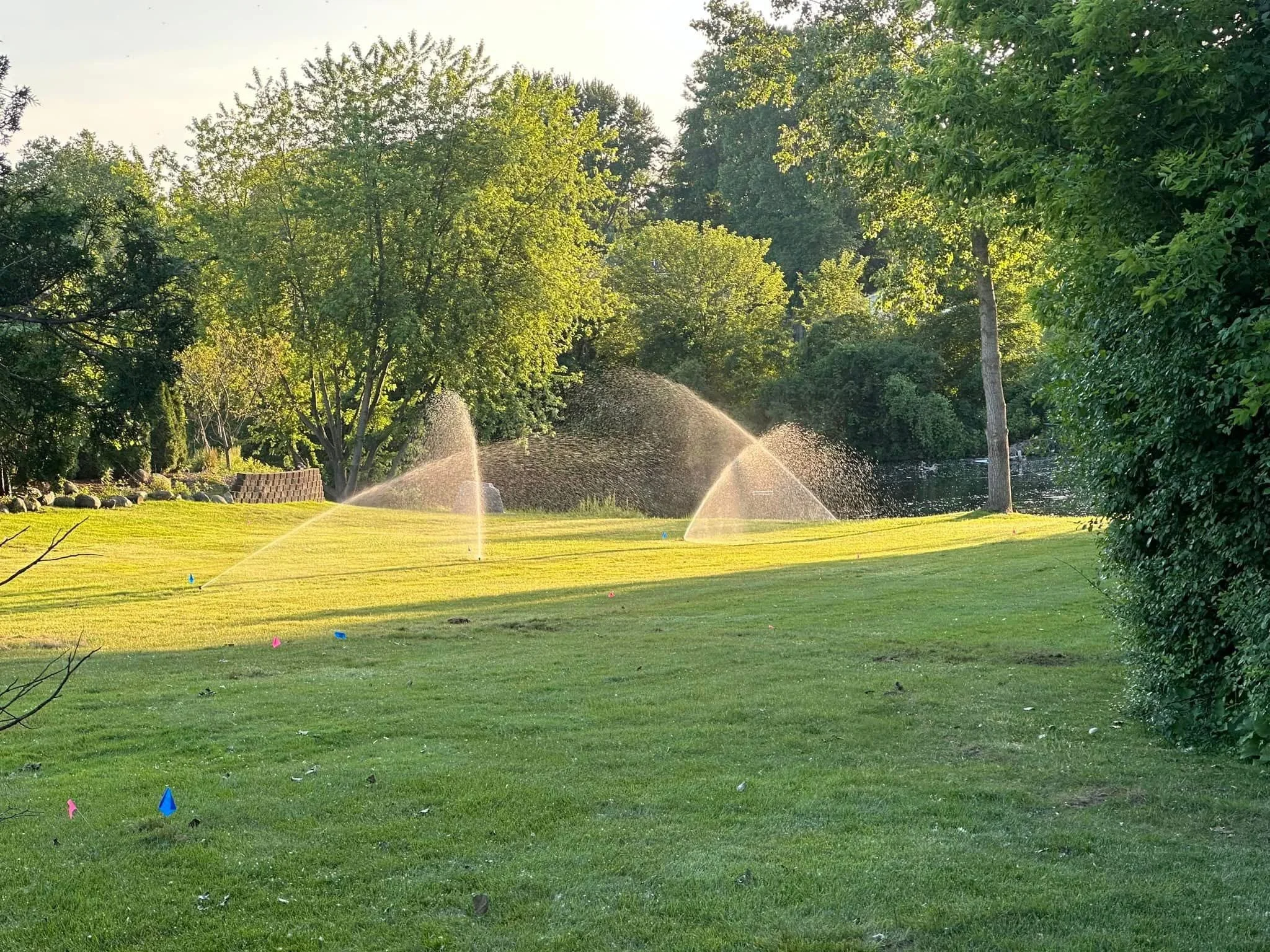A lush green park with trimmed grass, trees, and a pond with fountains in the background. There are colorful flags on the grass and a low brick wall on the left.