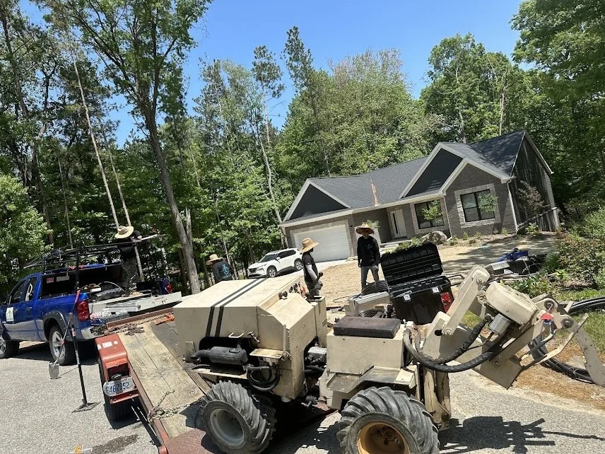 A group of people wearing wide-brimmed hats working outdoors near a modern house with a sloped roof surrounded by trees, with a blue utility truck and a piece of heavy machinery on a trailer.
