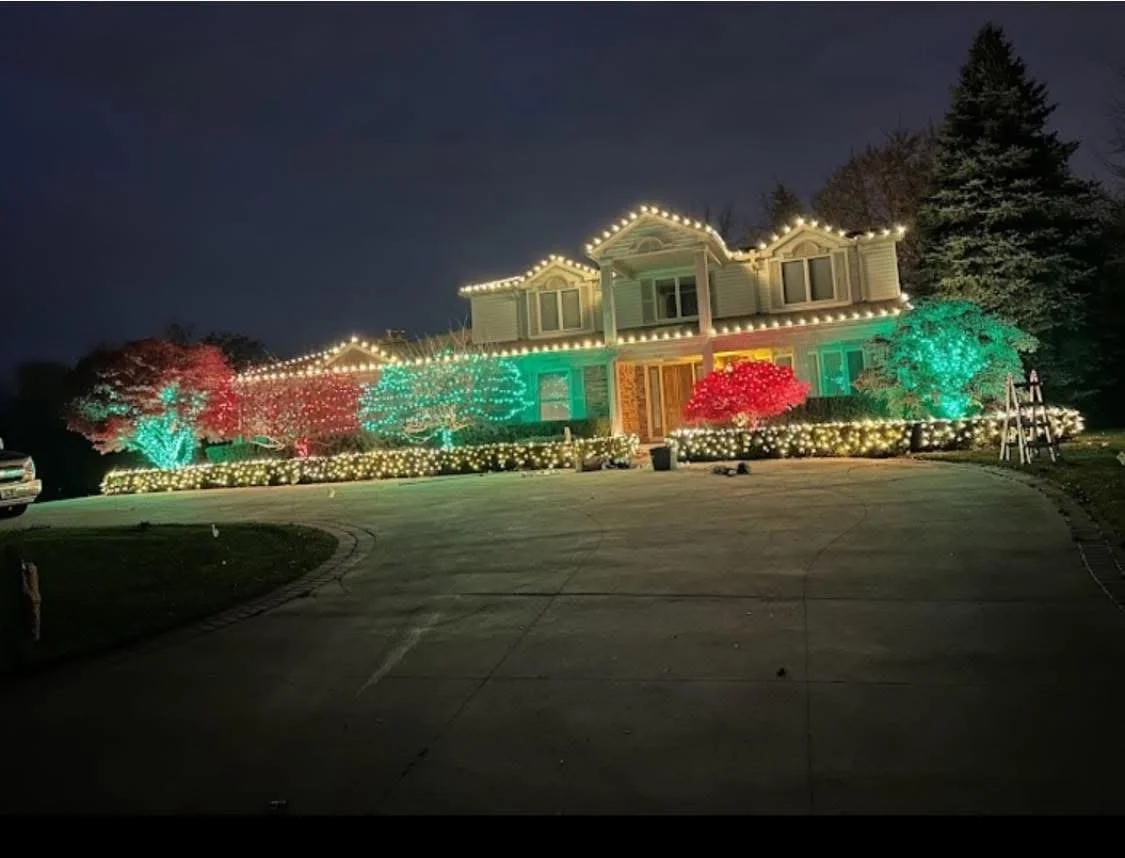 A house decorated with colorful Christmas lights, including red, green, and white, on trees and the roof, with a dark sky in the background.