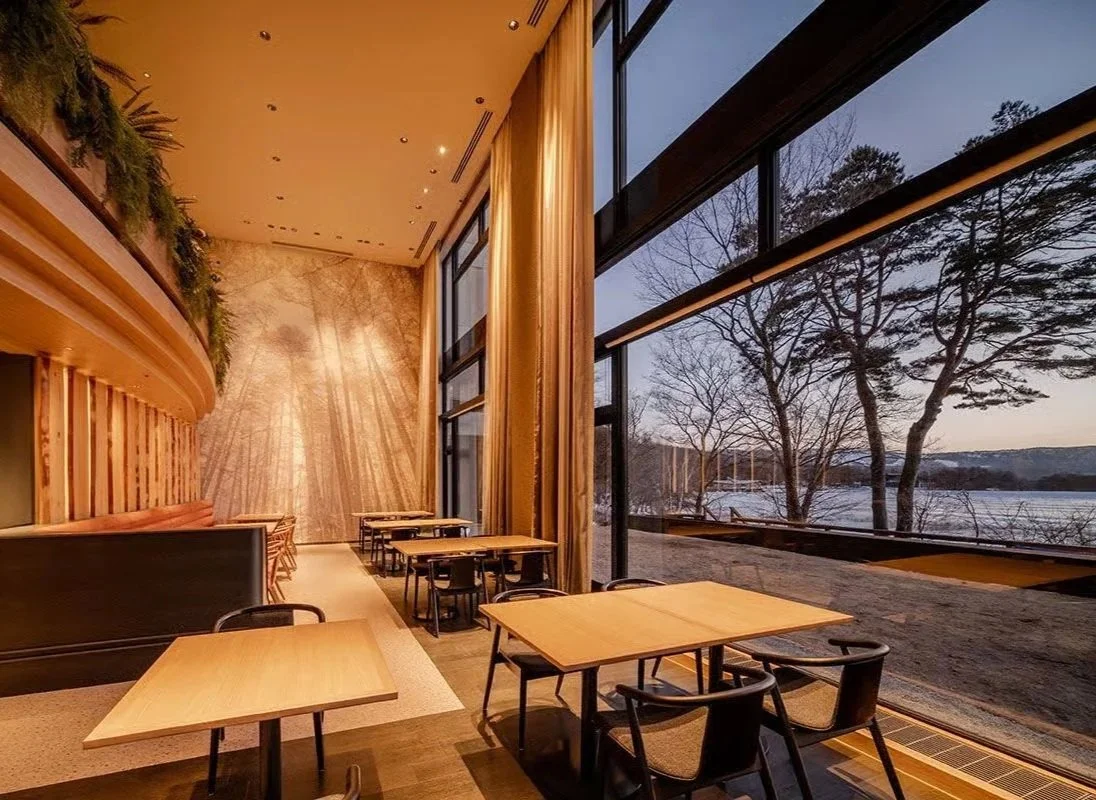 Interior of a modern restaurant with wooden tables and black chairs, large windows showing a lakeside view with leafless trees during dusk.