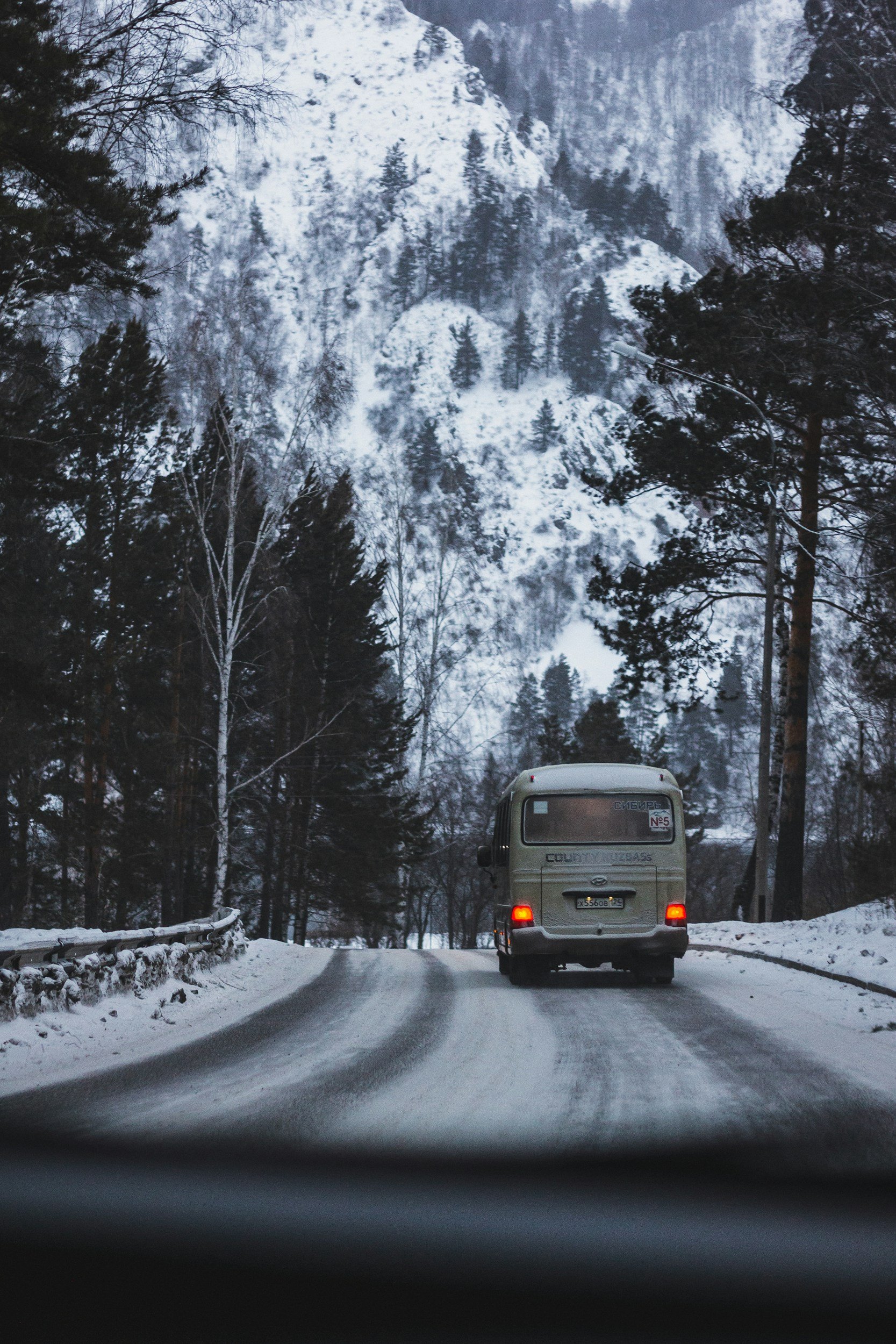 Snow-covered mountain landscape with trees, viewed through a car window on a snowy road, with a bus in front.