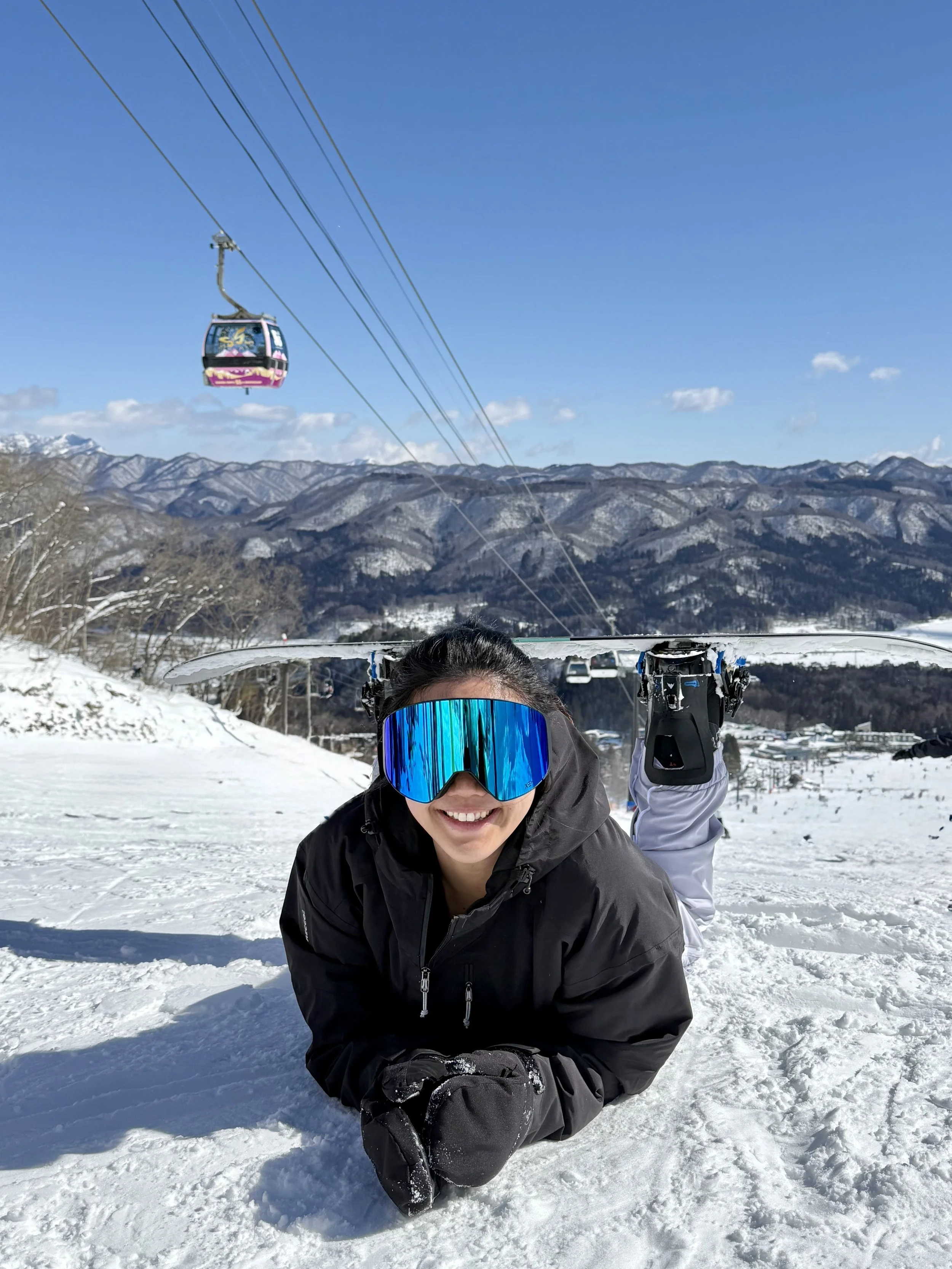 Person in ski goggles and winter gear smiling while kneeling in snow with snow-covered mountains and ski lift in the background.