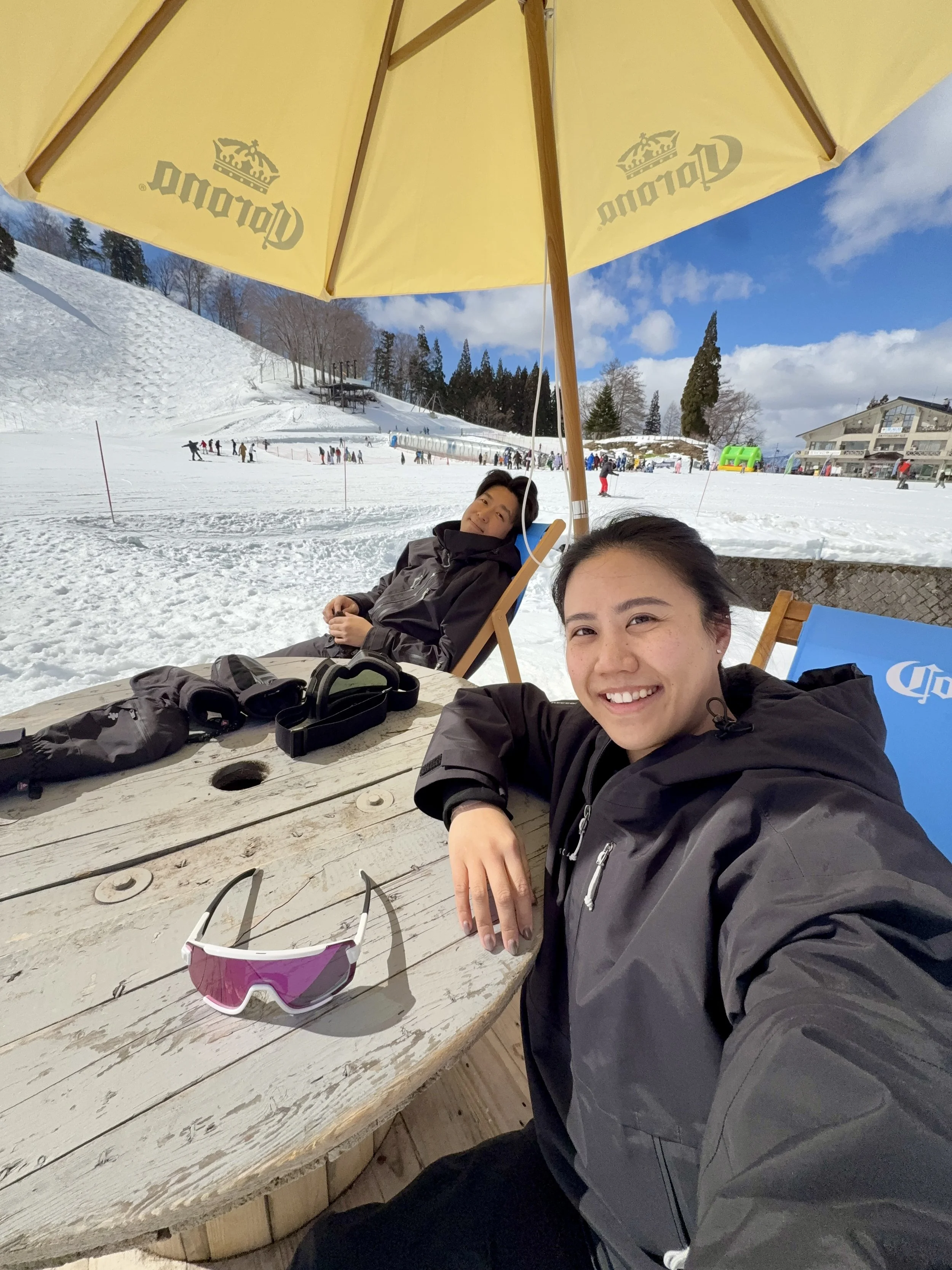Smiling woman taking a selfie at a snowy ski resort, with a man sitting behind her under a yellow umbrella, sunglasses on a wooden table, and skiers on the slope in the background.
