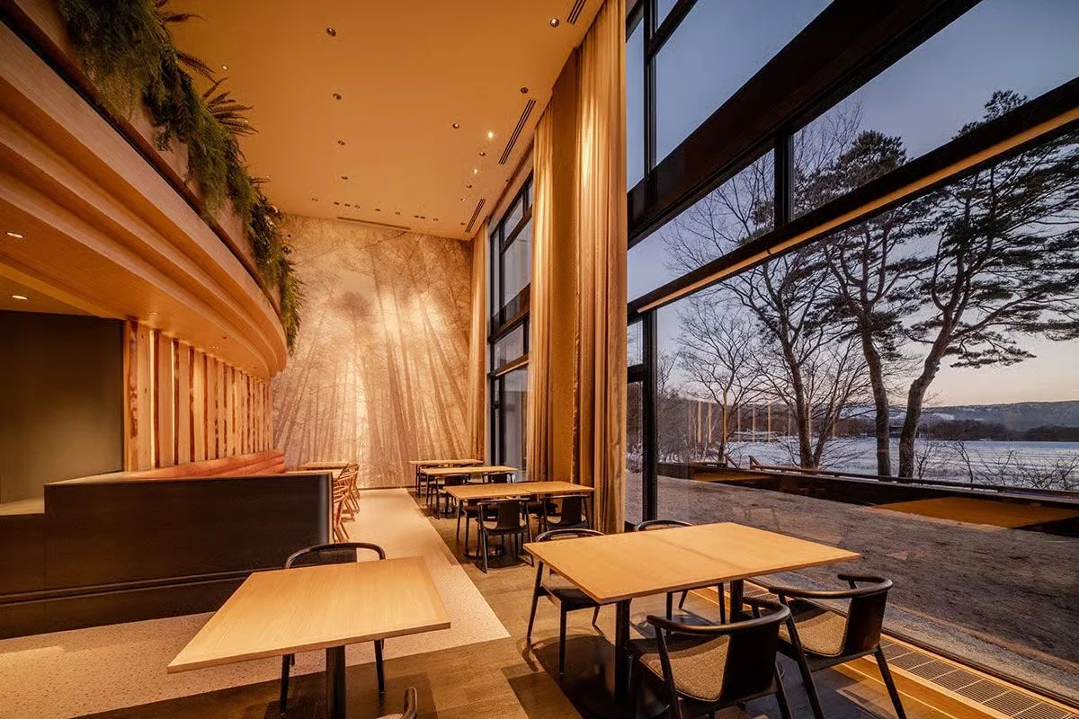 Interior of a restaurant with wooden tables and chairs, large floor-to-ceiling windows showing a view of trees and a river outside, elegant curtains, and warm lighting.