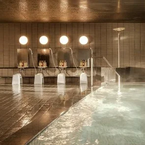 Indoor hot spring or spa with water jets, wooden deck, and tiled wall, illuminated by round overhead lights.
