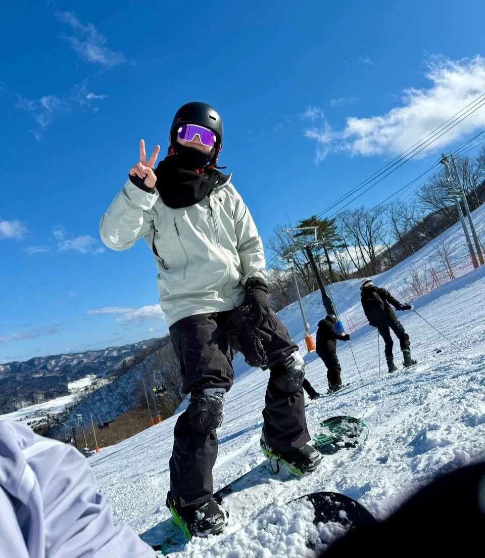 Person on a snowboard making a peace sign, dressed in winter gear with a helmet and ski goggles at a snowy ski resort, with others skiing in the background and mountains under a blue sky.