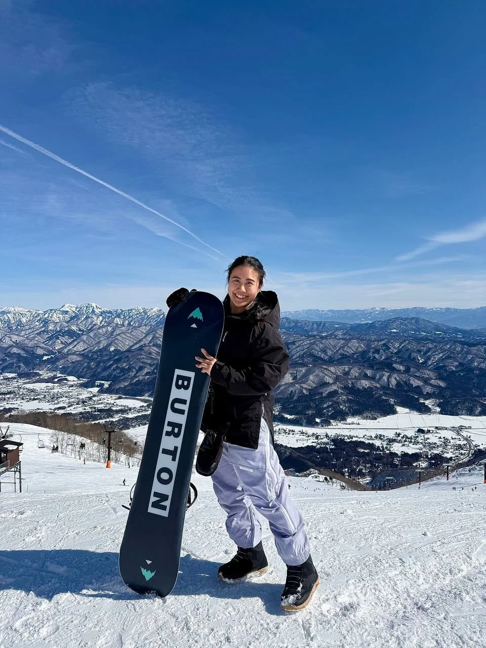 A smiling woman in winter gear standing on a snow-covered mountain slope, holding a snowboard with "BURTON" on it, with a snowy mountain landscape and blue sky in the background.