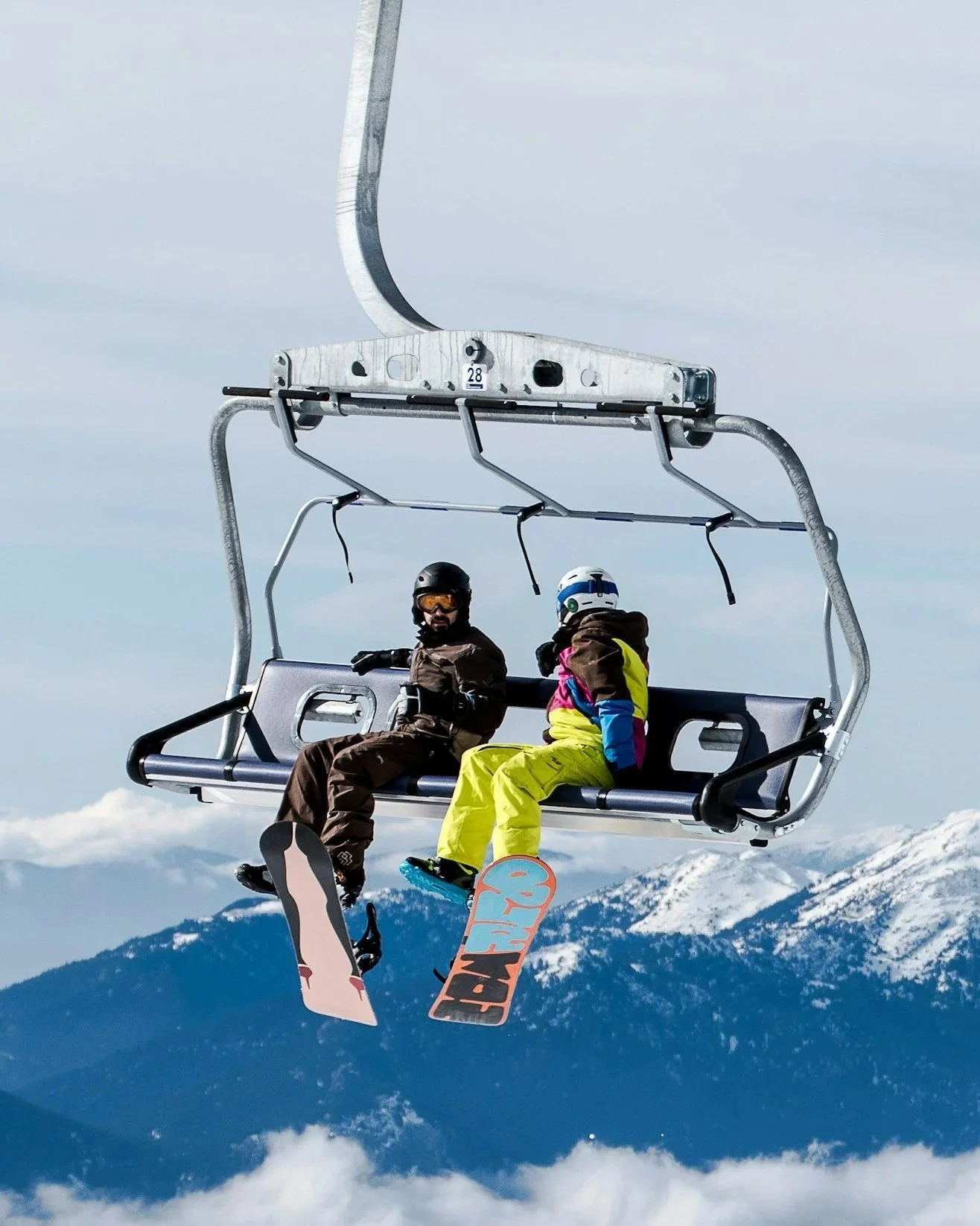 Two skiers sitting on a ski lift with snow-covered mountains in the background.