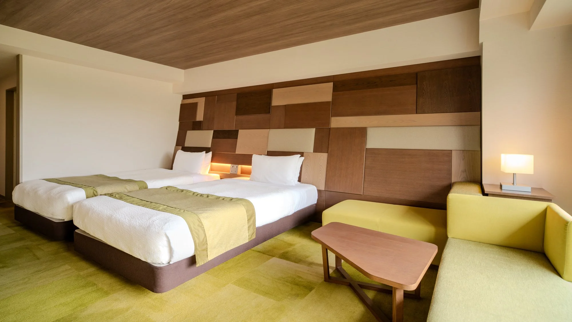 Hotel room with two beds, a built-in wooden headboard wall, a yellow armchair, a small wooden coffee table, and bedside lamps.