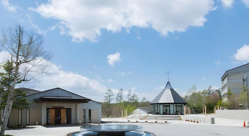 A church with a round glass structure and a cross on top and a modern building beside it, set in an open plaza with trees and a blue sky with clouds.