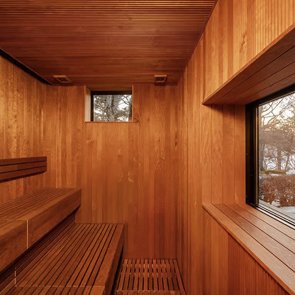Interior of a wooden sauna with benches on the left, a small window at the top, and a large window on the right showing trees outside.