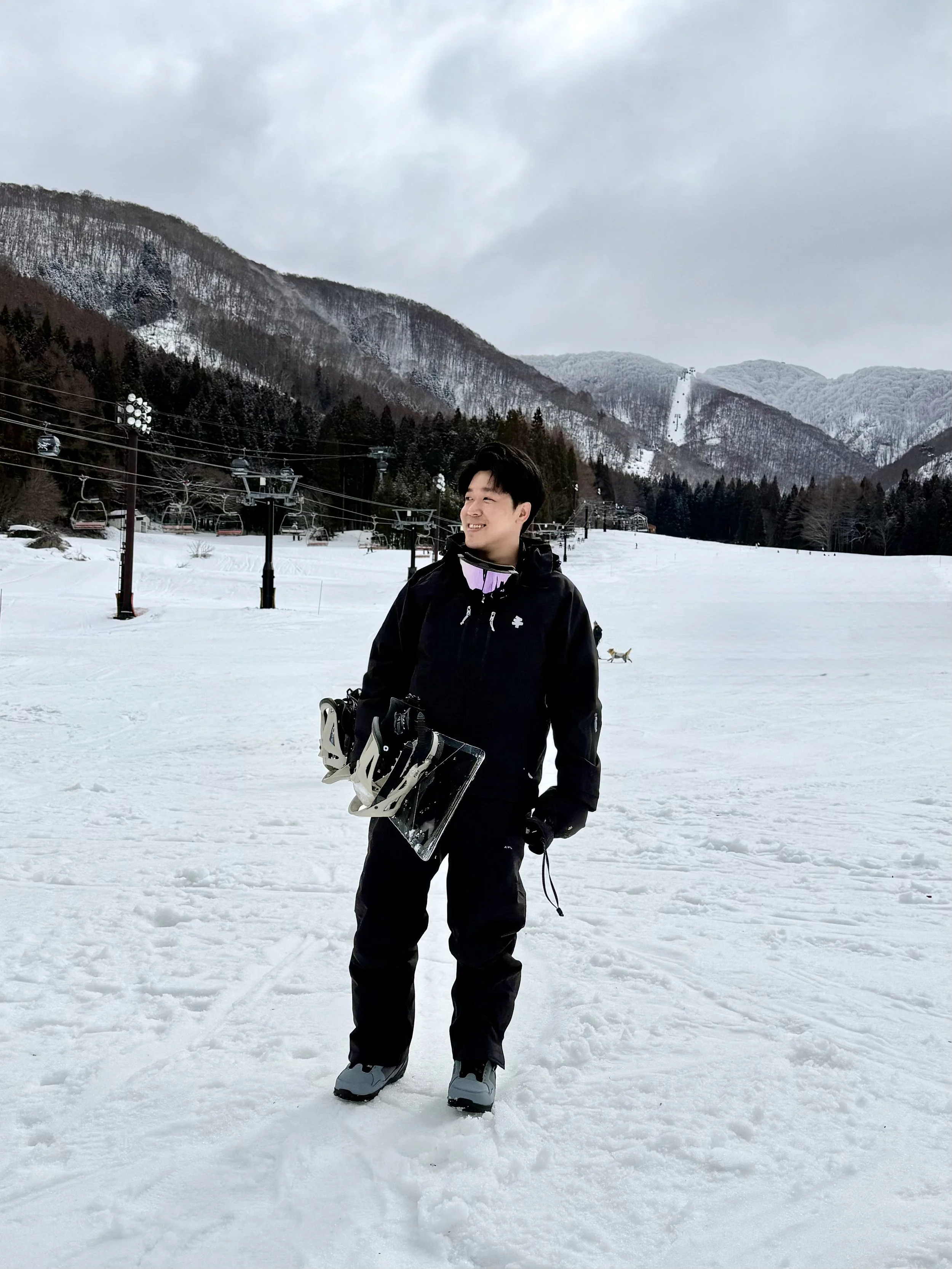 A young man in winter clothing is standing on snow with mountains in the background, holding snowboarding gear, smiling, with ski lifts and snow-covered trees visible.