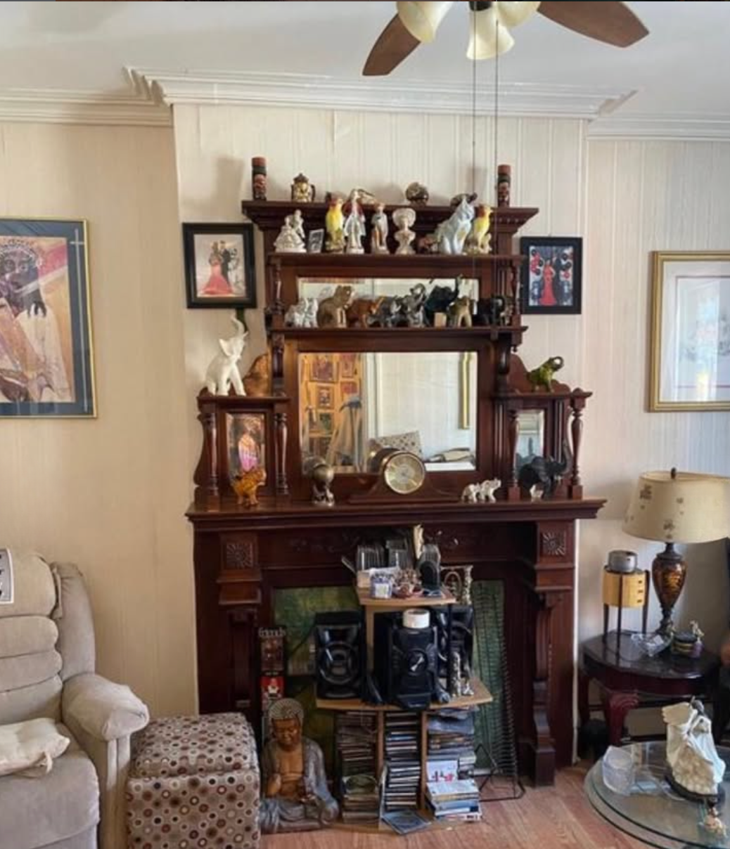A living room fireplace mantel decorated with numerous small statues and figurines, framed photographs, a clock, and various decorative objects. The mantel has a large mirror with a wooden frame and a built-in shelf with more decorations. To the left, a beige sofa and a ottoman are visible, and a floor lamp with a patterned shade sits on a side table to the right. The wall is adorned with framed artwork, and a ceiling fan with lights hangs overhead.