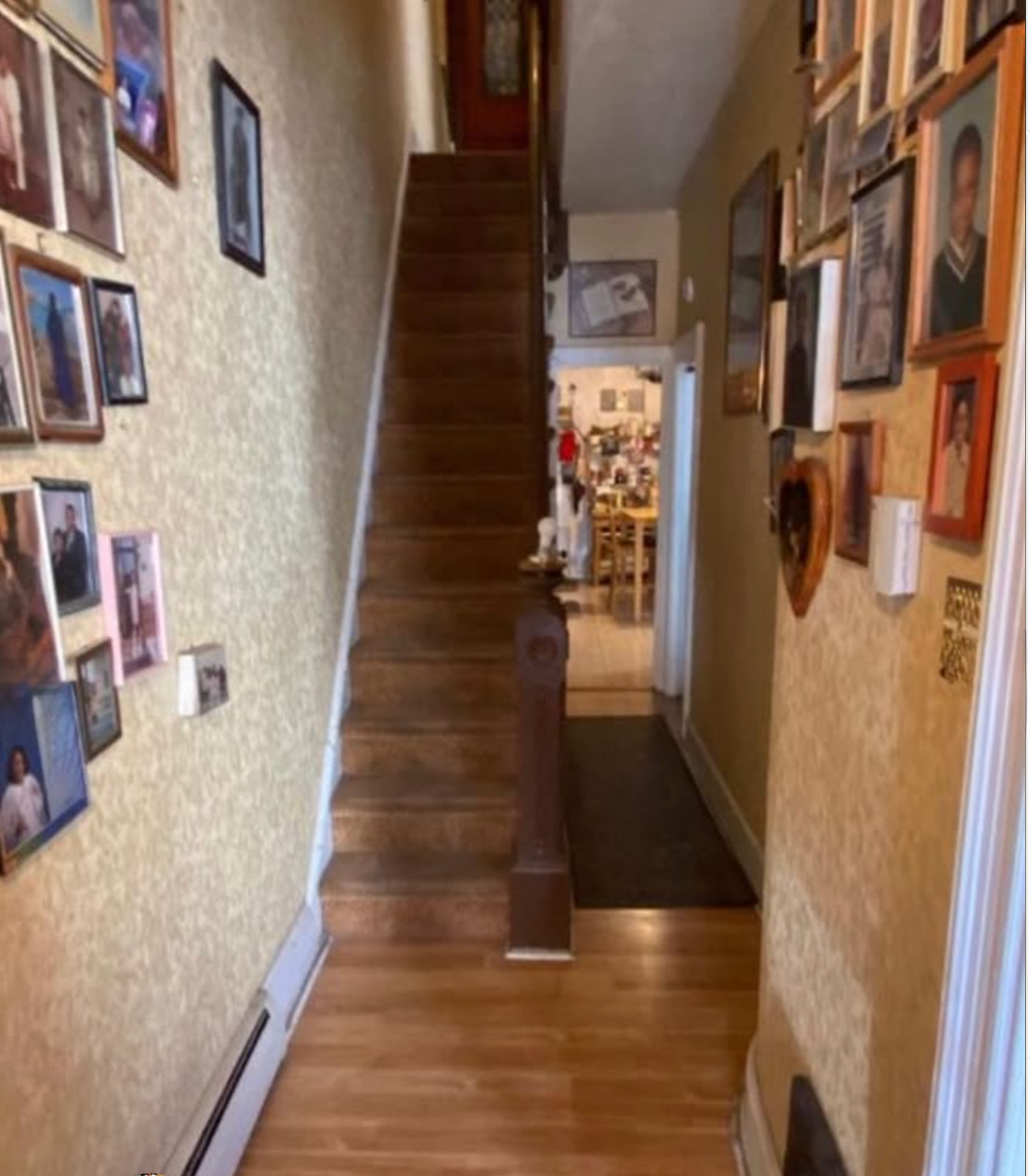 Hallway with stairs leading to upper floor, yellow patterned wallpaper, and framed photographs on both walls, leading to a kitchen area.