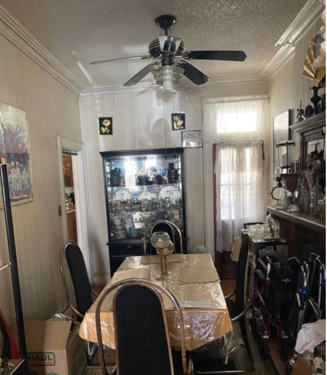 A dining room with a rectangular table covered in a yellow tablecloth and six black chairs. There is a glass cabinet filled with china and decorative items, and a ceiling fan with four blades. The room has two windows with white curtains, and various decorative items on shelves and walls.