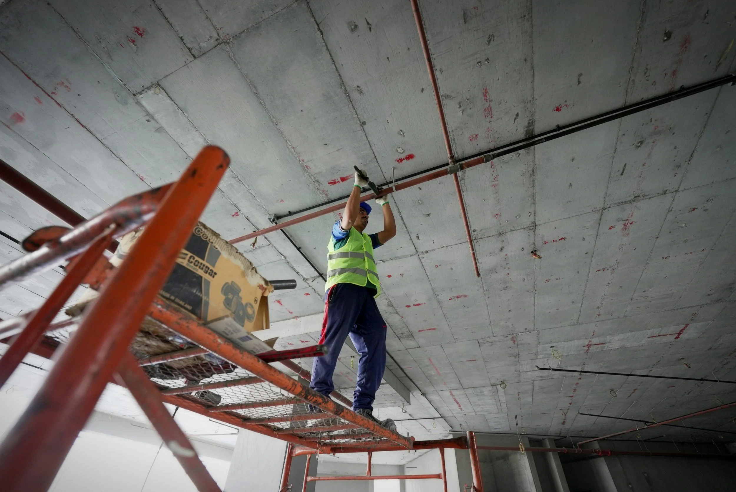 A construction worker in a reflective vest and gloves standing on scaffolding, working on the ceiling of a building with exposed pipes and electrical conduits.
