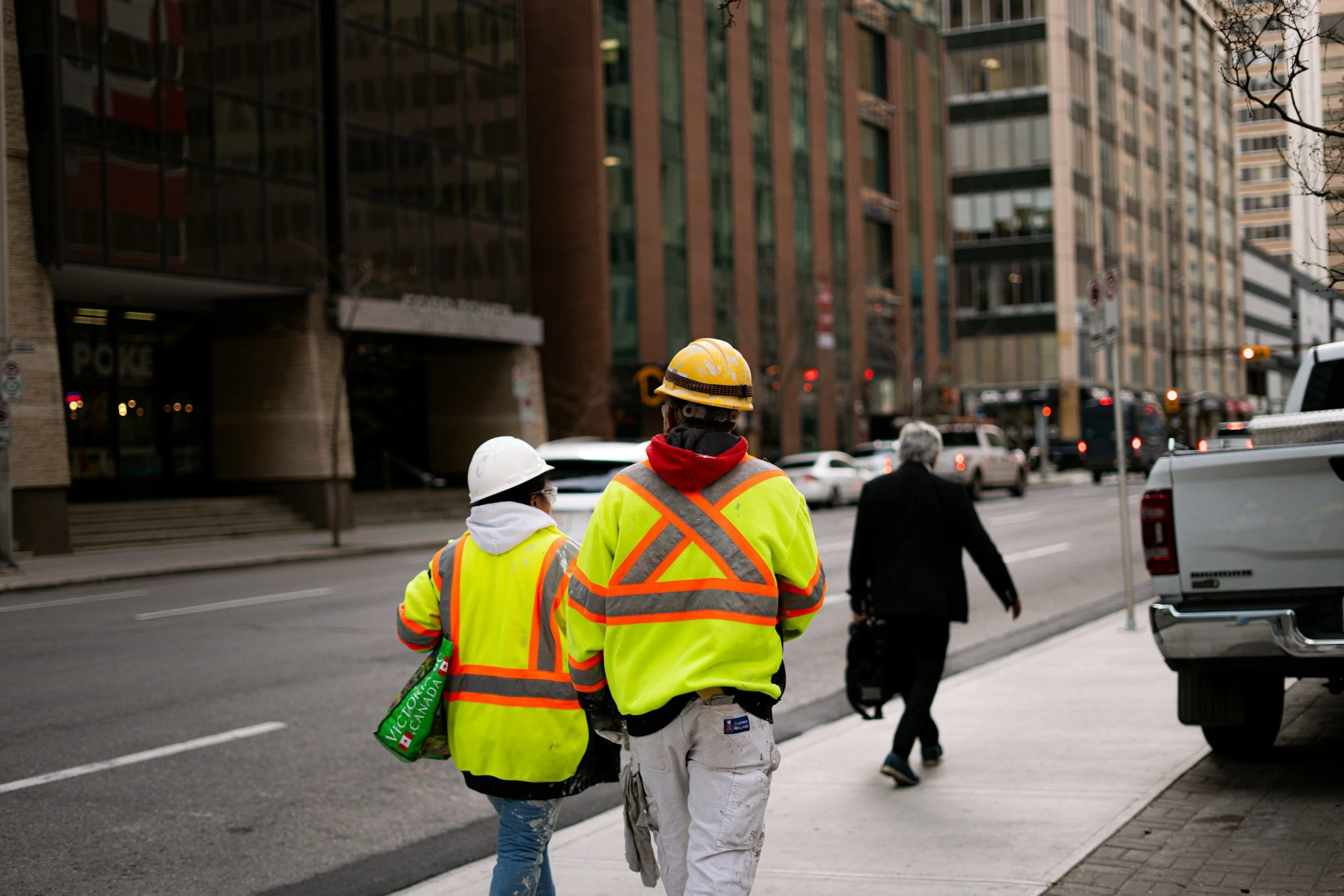 Two construction workers wearing yellow safety vests and helmets crossing the street in an urban area, with tall buildings and cars in the background.