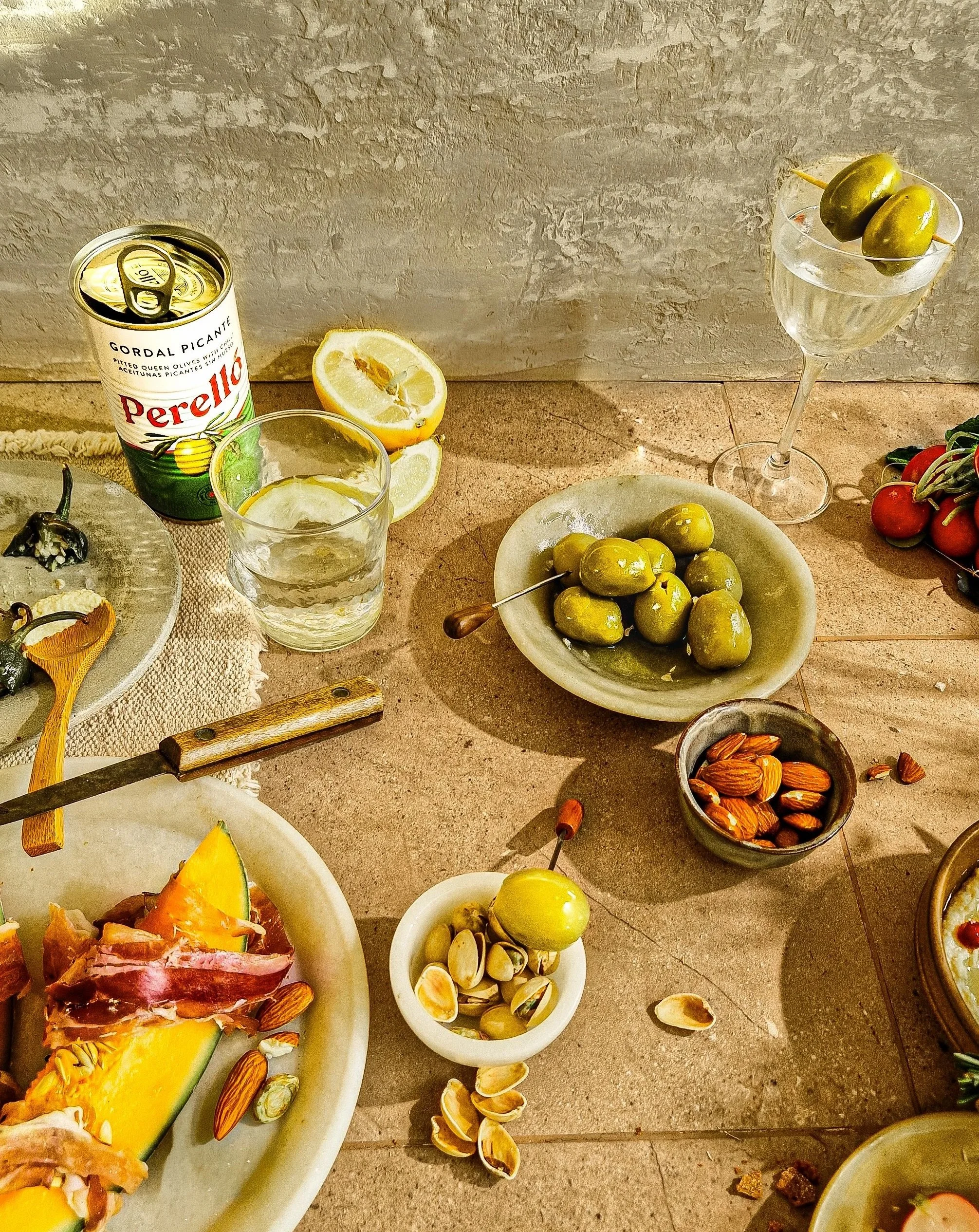 A variety of snacks on a table including a can of Perello pickled olives, a lemon, a glass of water, green olives on a skewer in a glass, a bowl of green olives, a bowl of almonds, and slices of cured meat with cheese and a slice of melon.