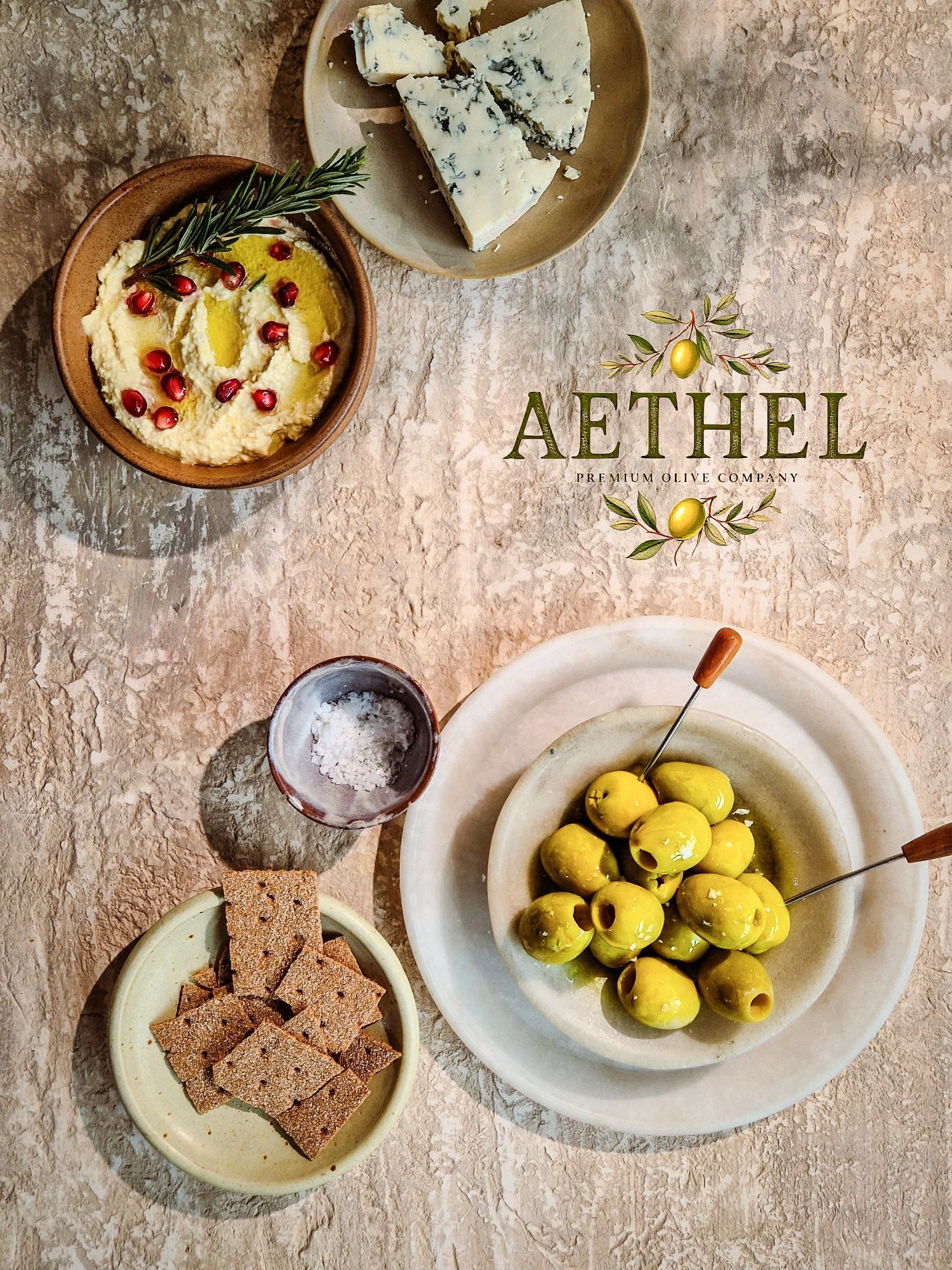 Various bowls of food including green olives, blue cheese, crackers, and a plate of hummus garnished with pomegranate seeds and rosemary, on a textured surface with a logo reading 'AETHEL Premium Olive Company'.