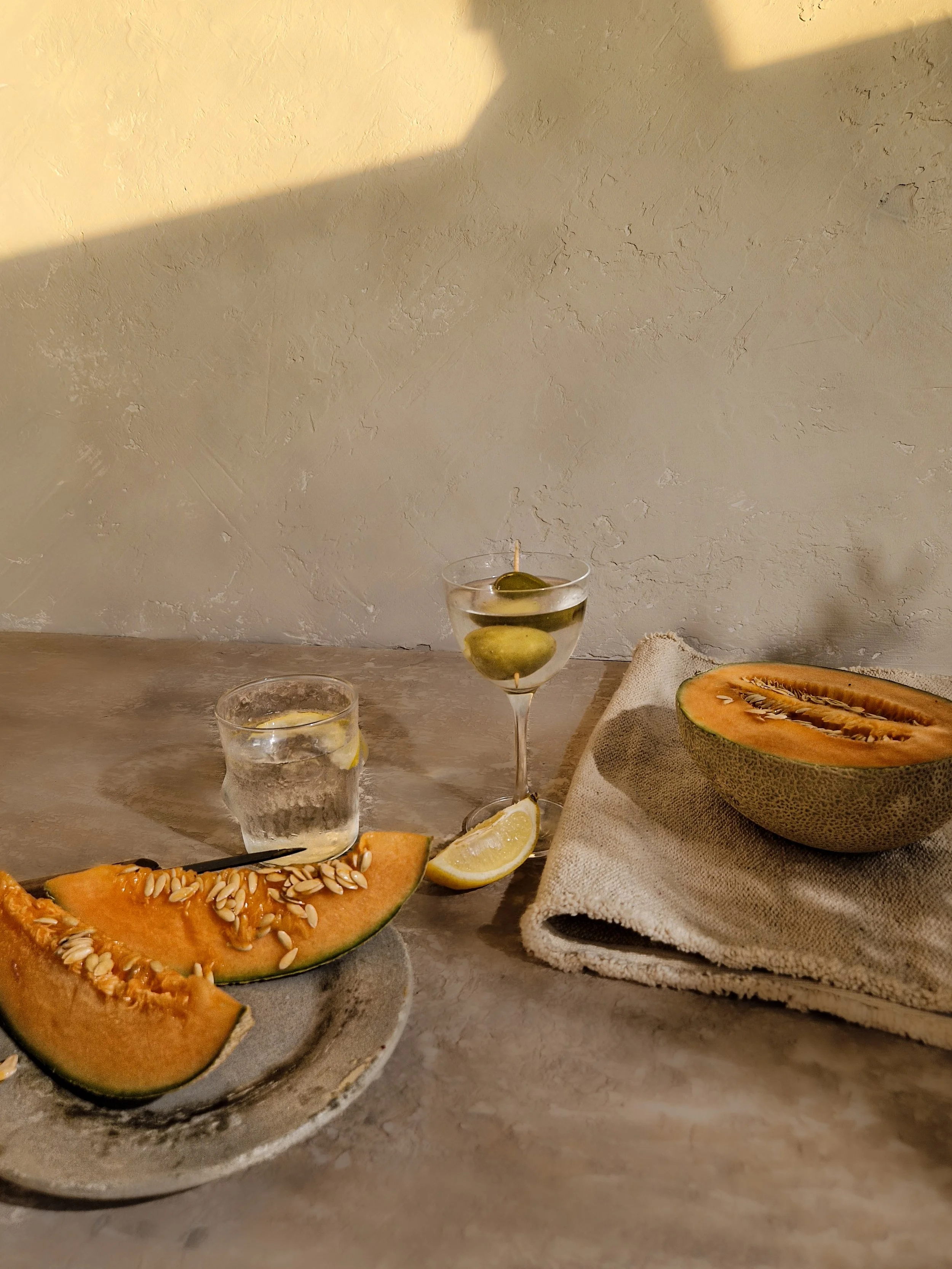 Halved cantaloupe on a ceramic plate, a glass of infused water with lemon and olives, a glass of water with lemon slices, and a halved lemon on a cloth napkin on a textured beige surface against a beige wall.