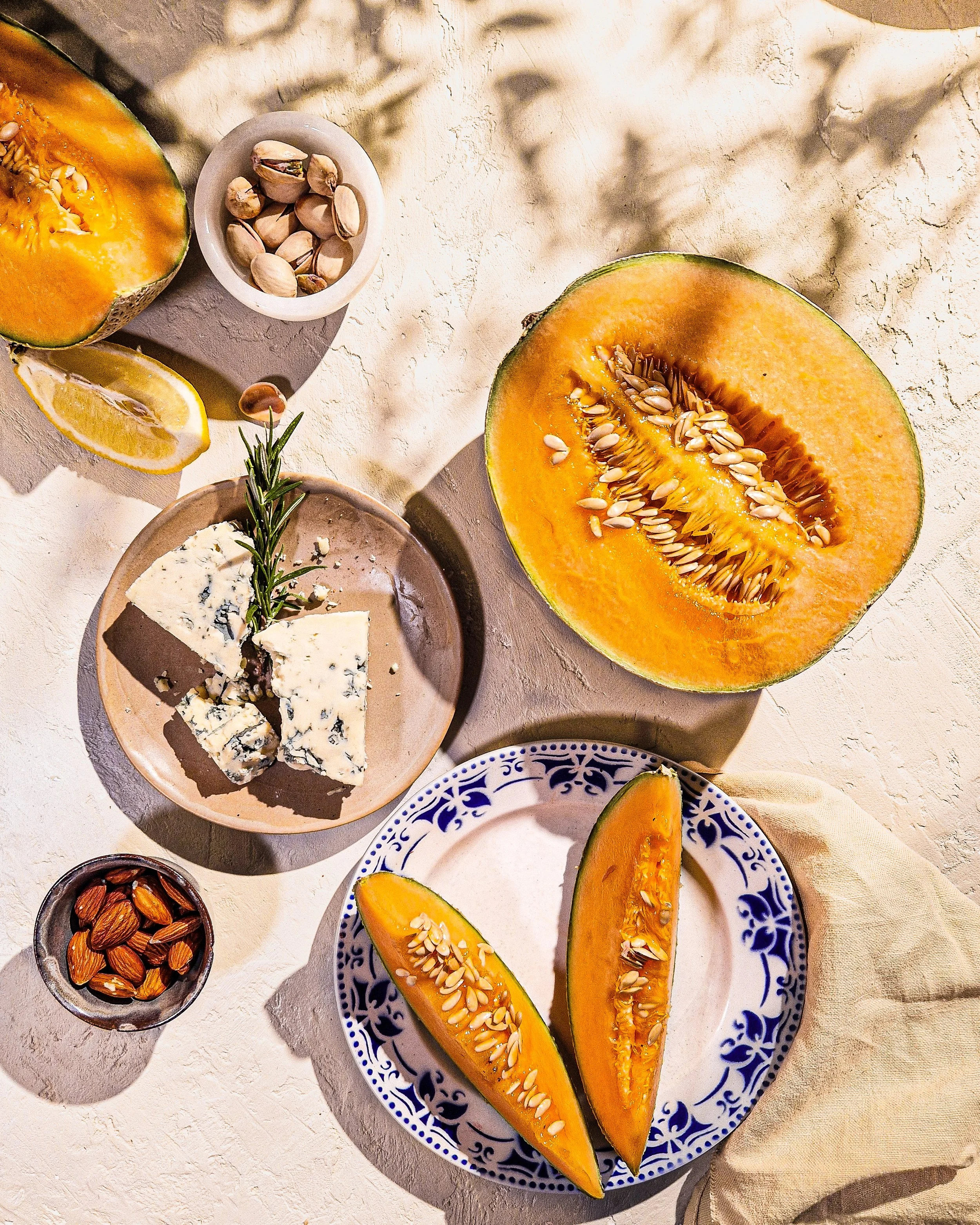 A sliced orange and two halves of a cantaloupe melon on a decorative blue and white plate, with a small bowl of pistachios, almonds, and a wedge of lemon in the background.