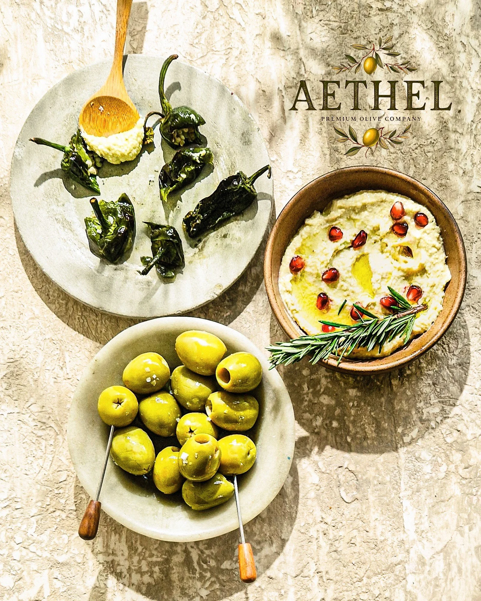 Green olives on skewers, a bowl of fresh yellow-green olives, a bowl of creamy white dip with pomegranate seeds, and a sprig of rosemary, set on a textured surface.