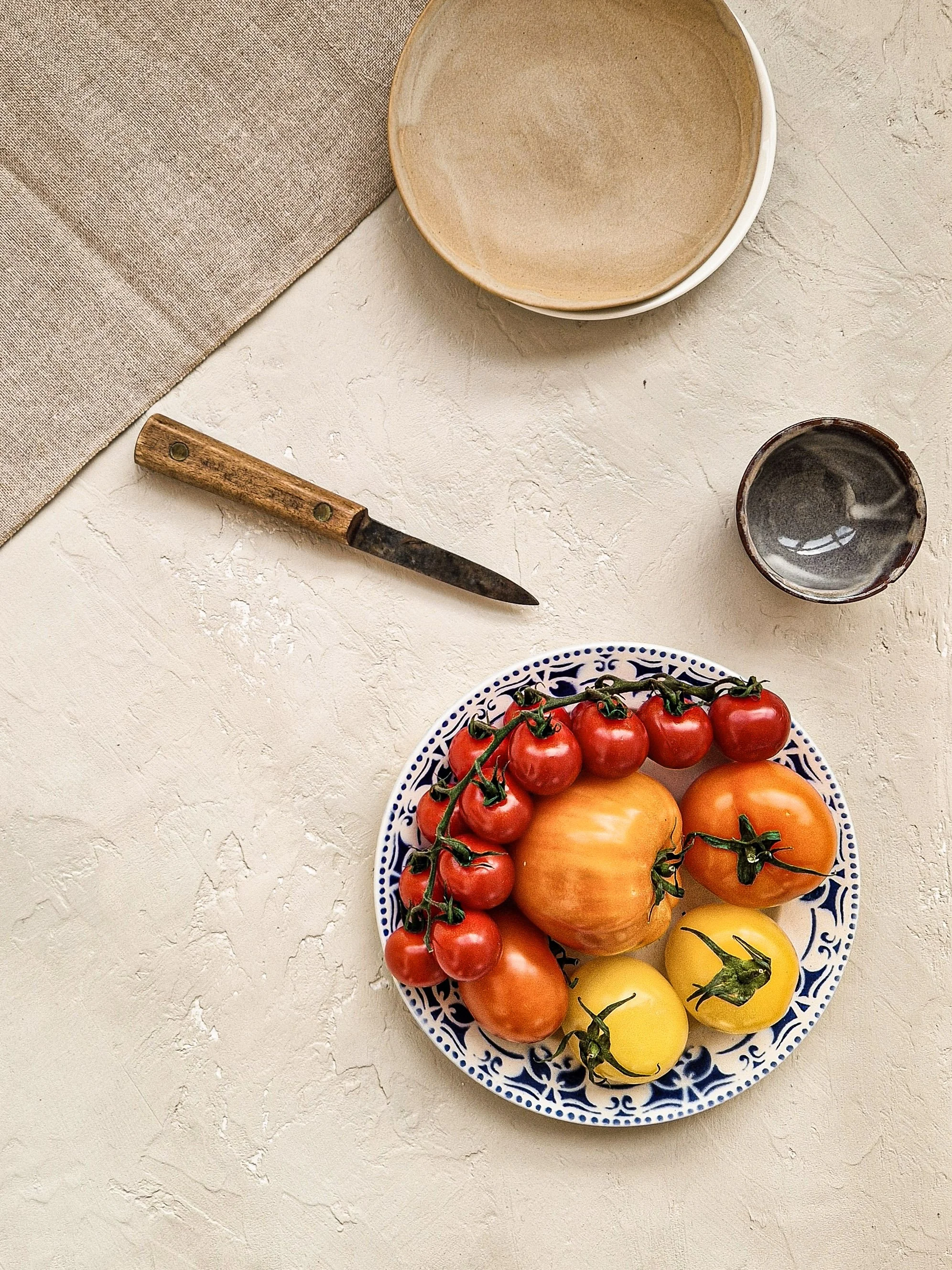 A white textured surface with a blue and white patterned plate of colorful tomatoes and peppers, and a small cup of water, along with a beige bowl, a wooden-handled knife, and a beige cloth in the background.