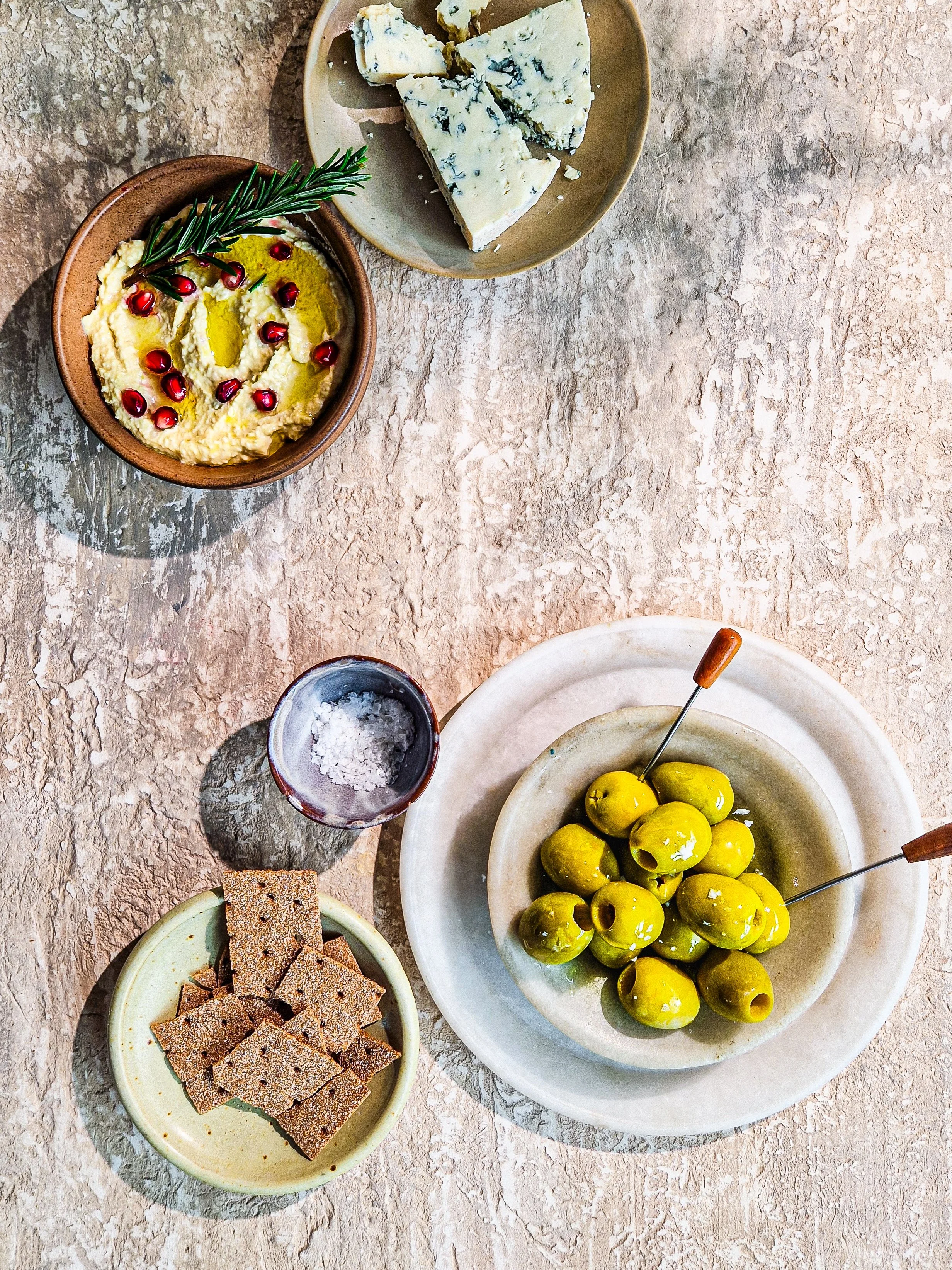 A table with small bowls containing various snacks: a bowl of green olives with a toothpick, a bowl of sliced bread or crackers, a bowl of yellow hummus topped with pomegranate seeds and a sprig of rosemary, a plate of blue cheese wedges, and a small container of coarse sea salt.