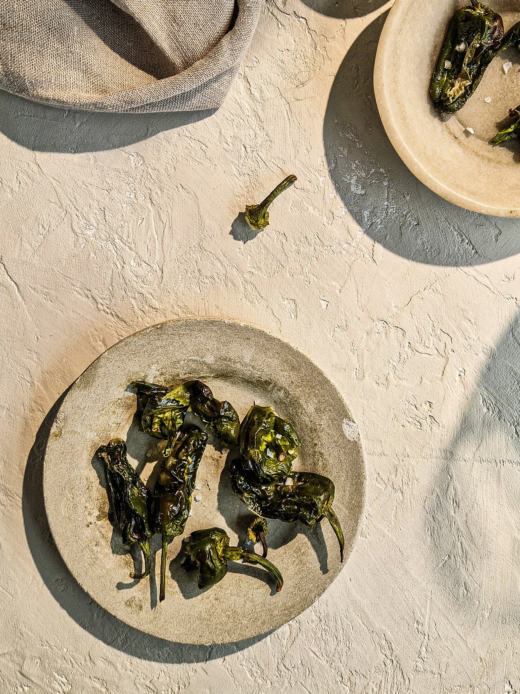 Several charred green peppers on white ceramic plates with a beige textured surface background.