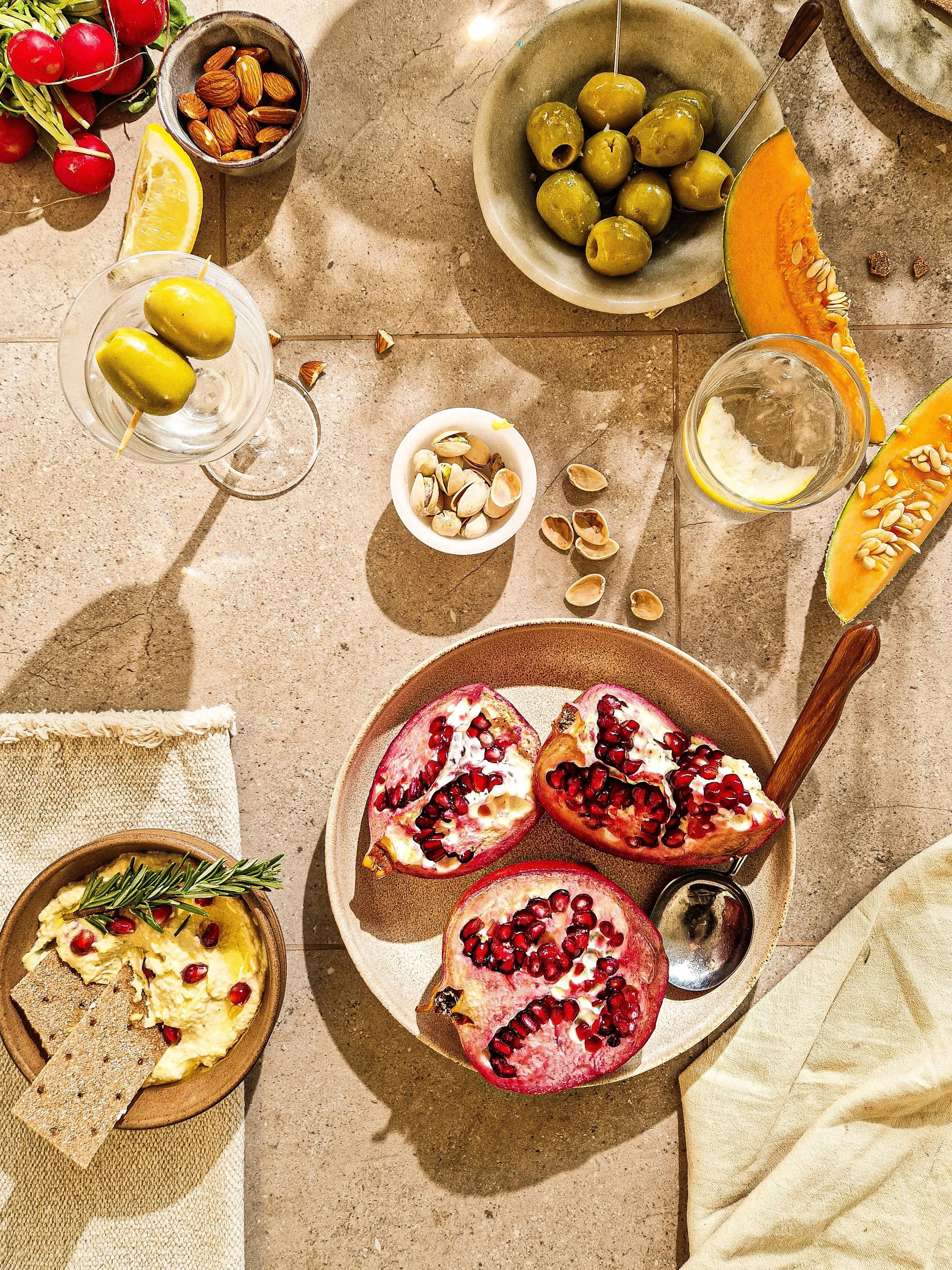 A flat lay of a Mediterranean-inspired spread featuring sliced pomegranate, green olives, roasted almonds, radishes, garlic, herbs, and lemon slices on a tiled surface.