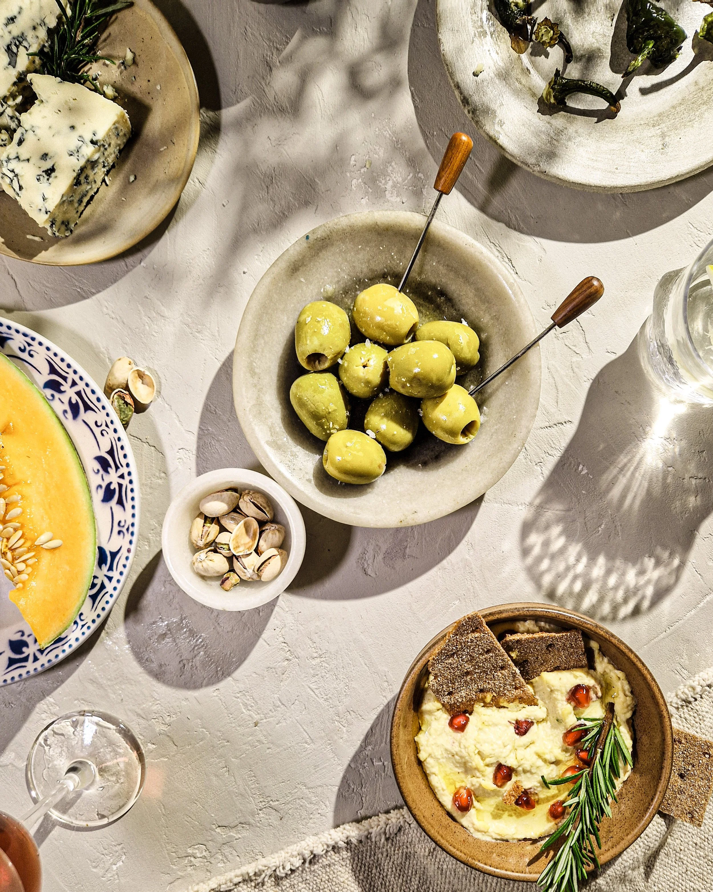 A table with various dishes including a bowl of green olives with skewers, a wedge of blue cheese with rosemary on a plate, a bowl of cracked pistachios, a slice of melons, and a bowl of creamy dip garnished with pomegranate seeds and rosemary. There is also an empty glass and a wooden bowl of dip with bread or crackers.