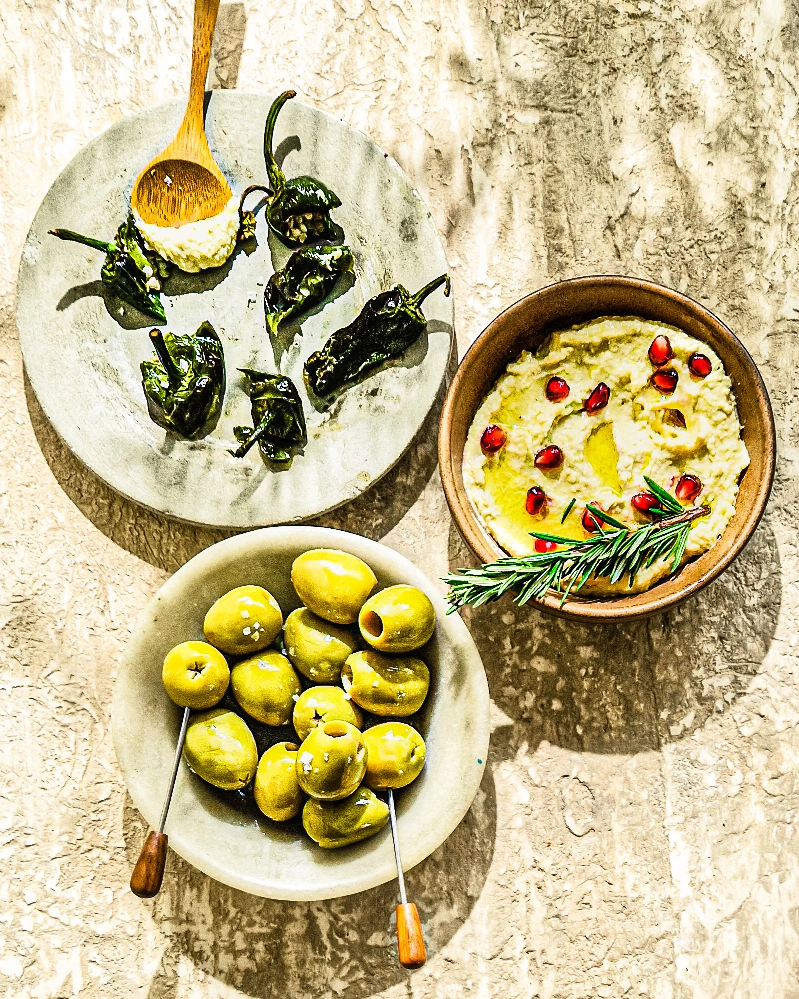 A plate of grilled green chili peppers with a wooden spoon of garlic mashed with salt, a bowl of yellow cherry tomatoes, and a bowl of creamy cheese garnished with pomegranate seeds and rosemary.