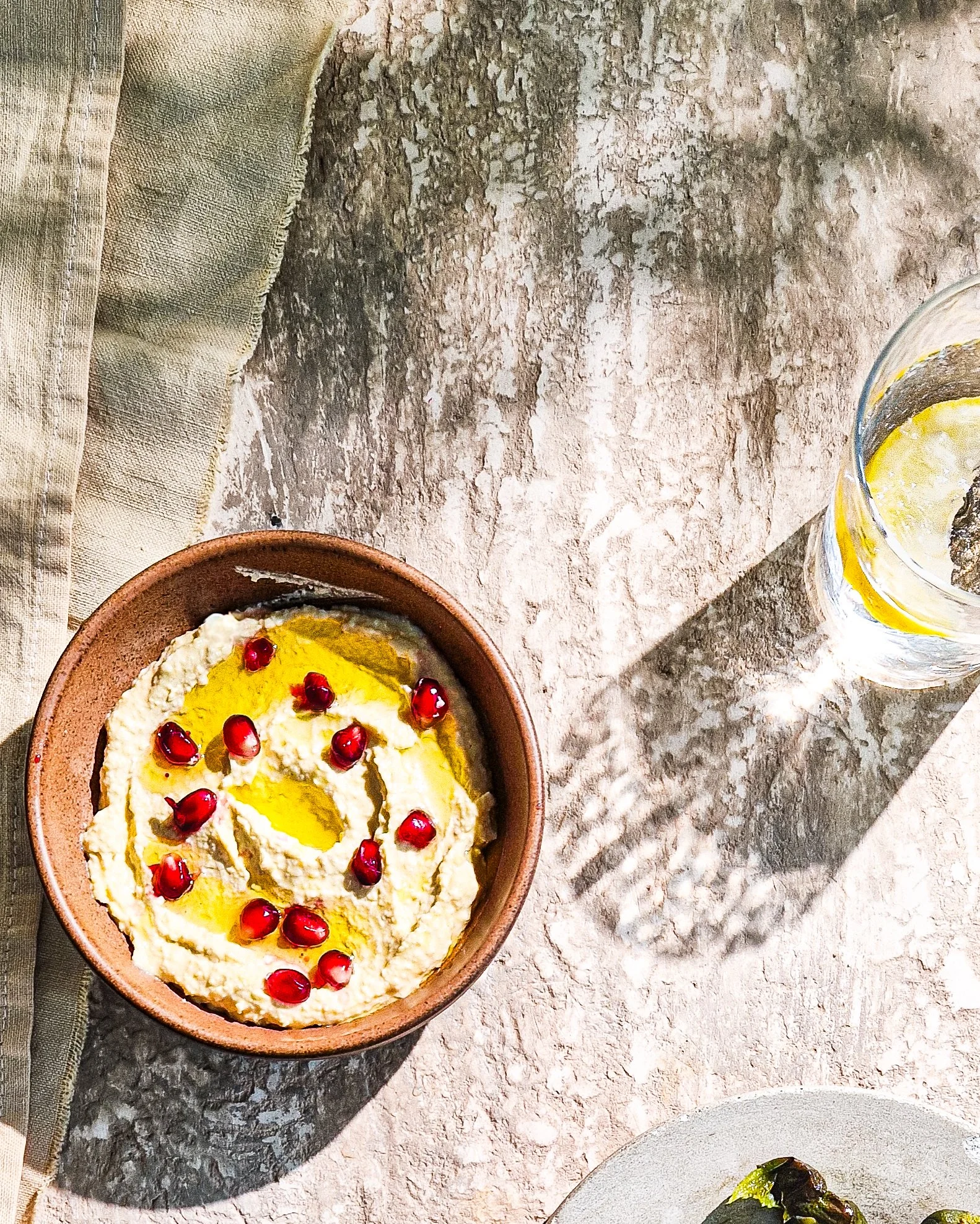 A terracotta bowl filled with Greek yogurt topped with pomegranate seeds and a drizzle of olive oil, placed on a rustic stone surface. To the right, a glass of water with lemon slices is partially visible. The background includes a linen napkin and a textured cloth.