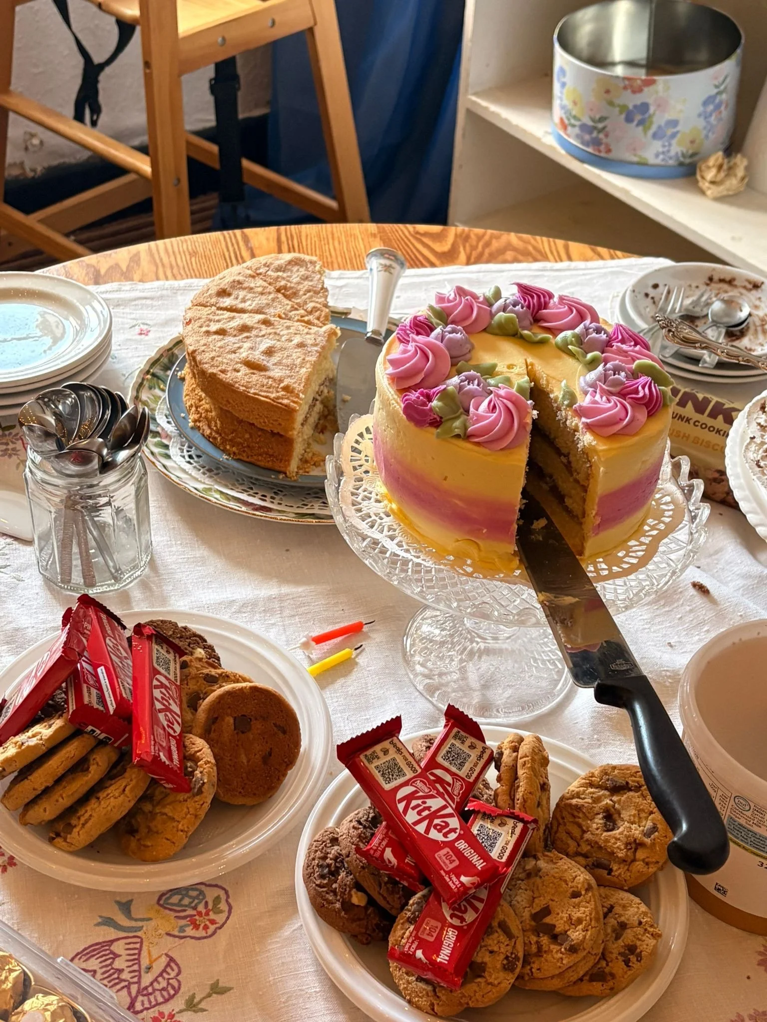 A table with various desserts including a decorated cake with pink and purple frosting, a partially sliced cake, cookies, and KitKat chocolate bars. There are plates, utensils, and a glass jar of spoons on the table.