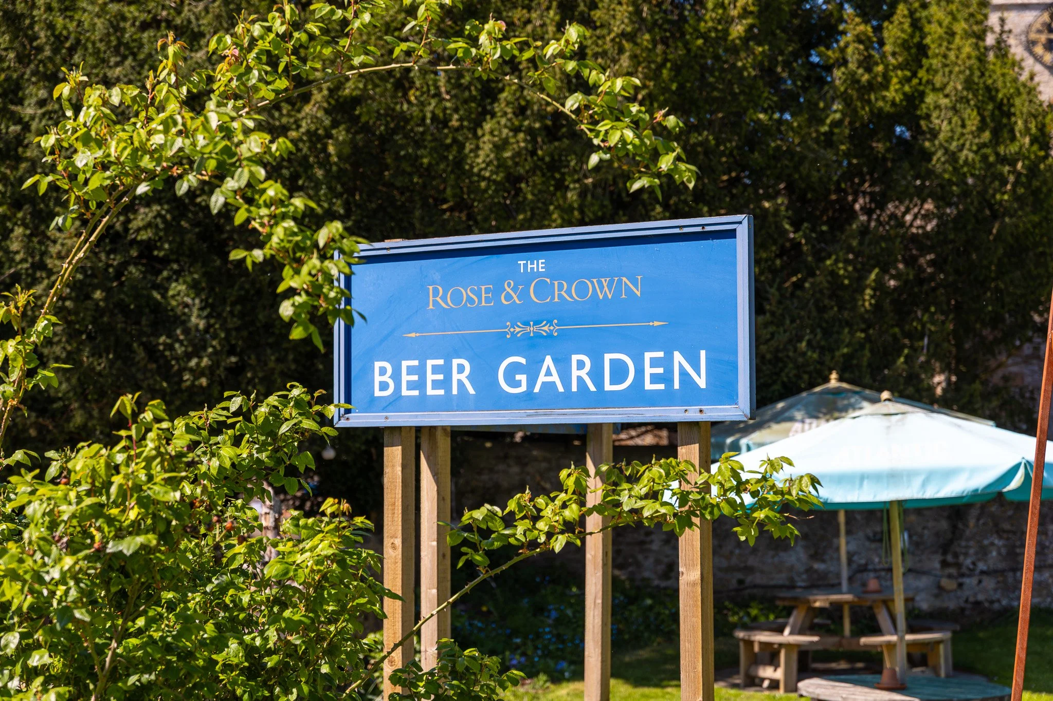 A blue sign that reads 'The Rose & Crown Beer Garden' is outdoors, surrounded by green foliage, with patio umbrellas and seating visible in the background.