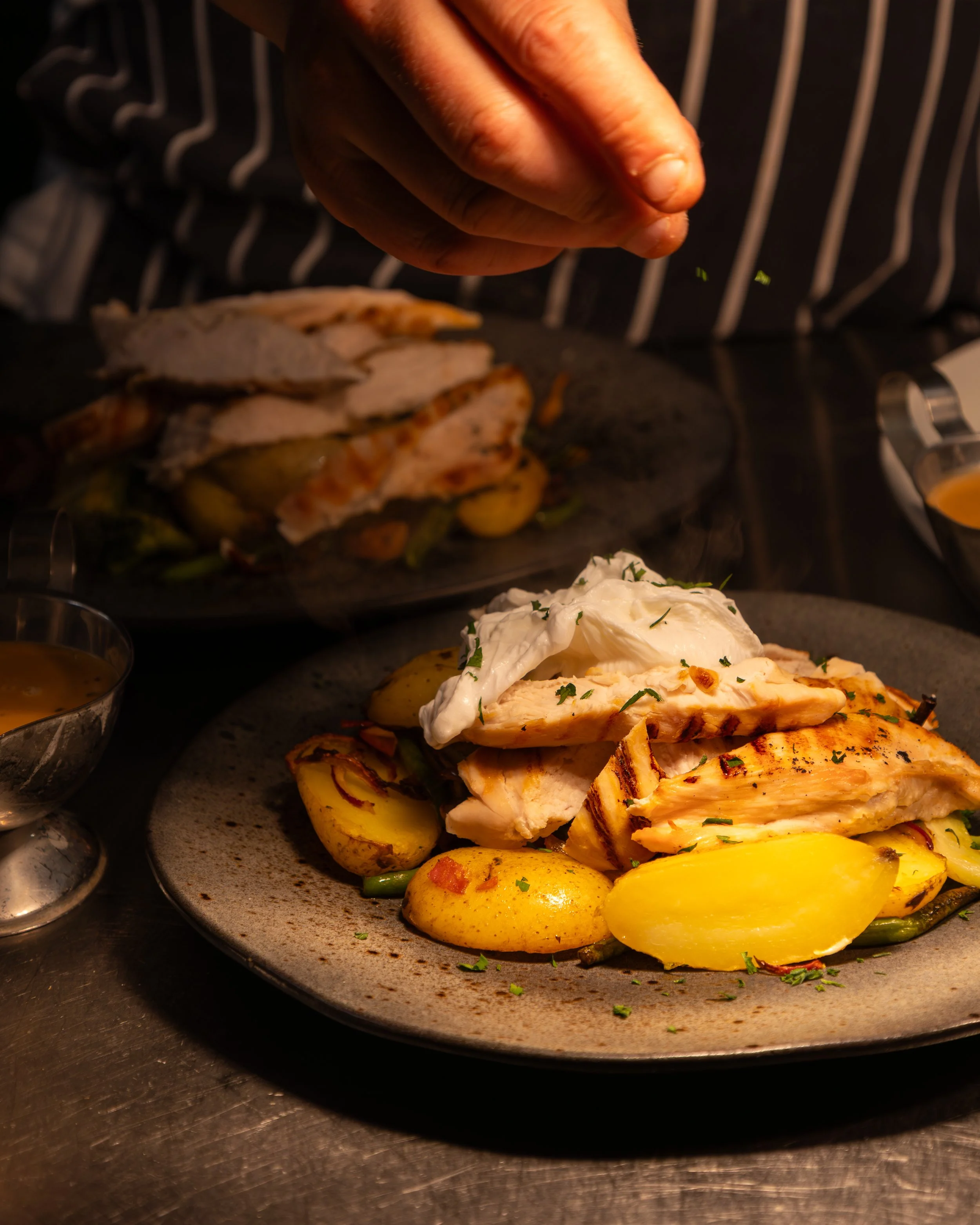 Close-up of a chef seasoning grilled chicken breast topped with herbs, served with roasted potatoes and green beans on a gray plate in a restaurant kitchen.