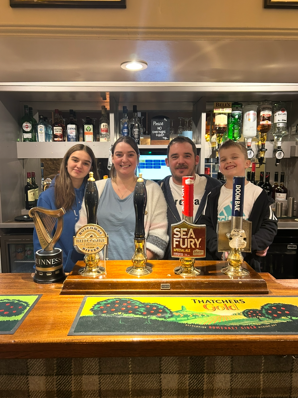 Four people standing behind a bar counter with beer taps, smiling for the photo. The bar has various bottles of alcohol in the background, and colorful mats on the counter feature green trees and the text "Thatchers Gold."