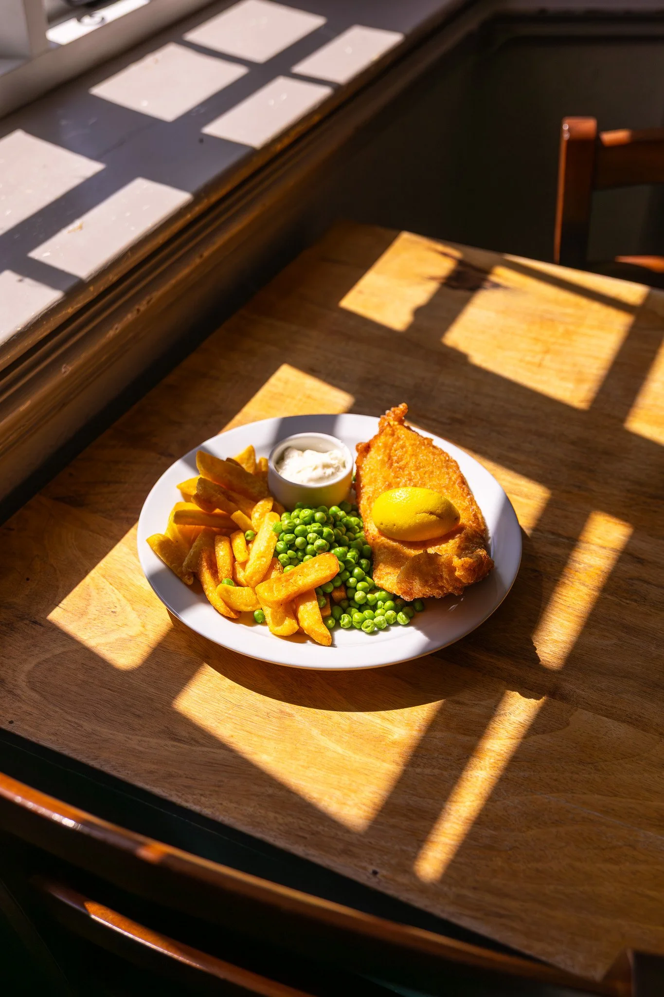 A plate of fish and chips with green peas, French fries with ketchup, a small cup of tartar sauce, and a lemon wedge, sitting on a wooden table with sunlight and shadows.