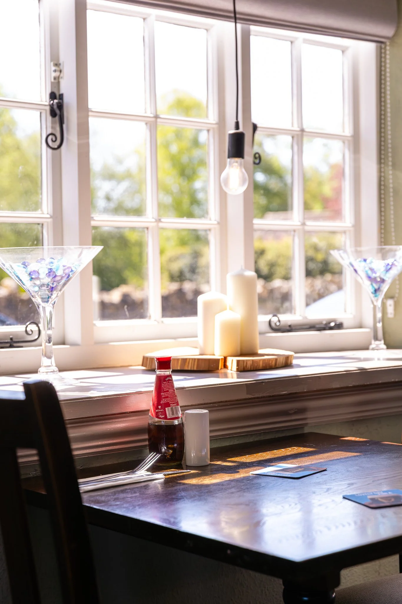 A kitchen window with views of green trees outside. On the windowsill, there are white candles, two glass vases filled with colorful decorative stones, a bottle of tomato ketchup, napkins, and a menu card. A black table is partially visible in the fo