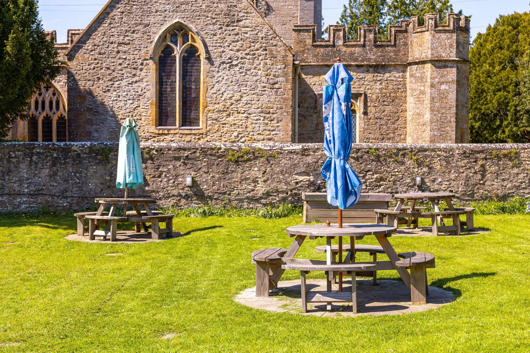 A stone church with Gothic windows, surrounded by a grassy yard with several picnic tables and umbrellas, and some trees in the background.