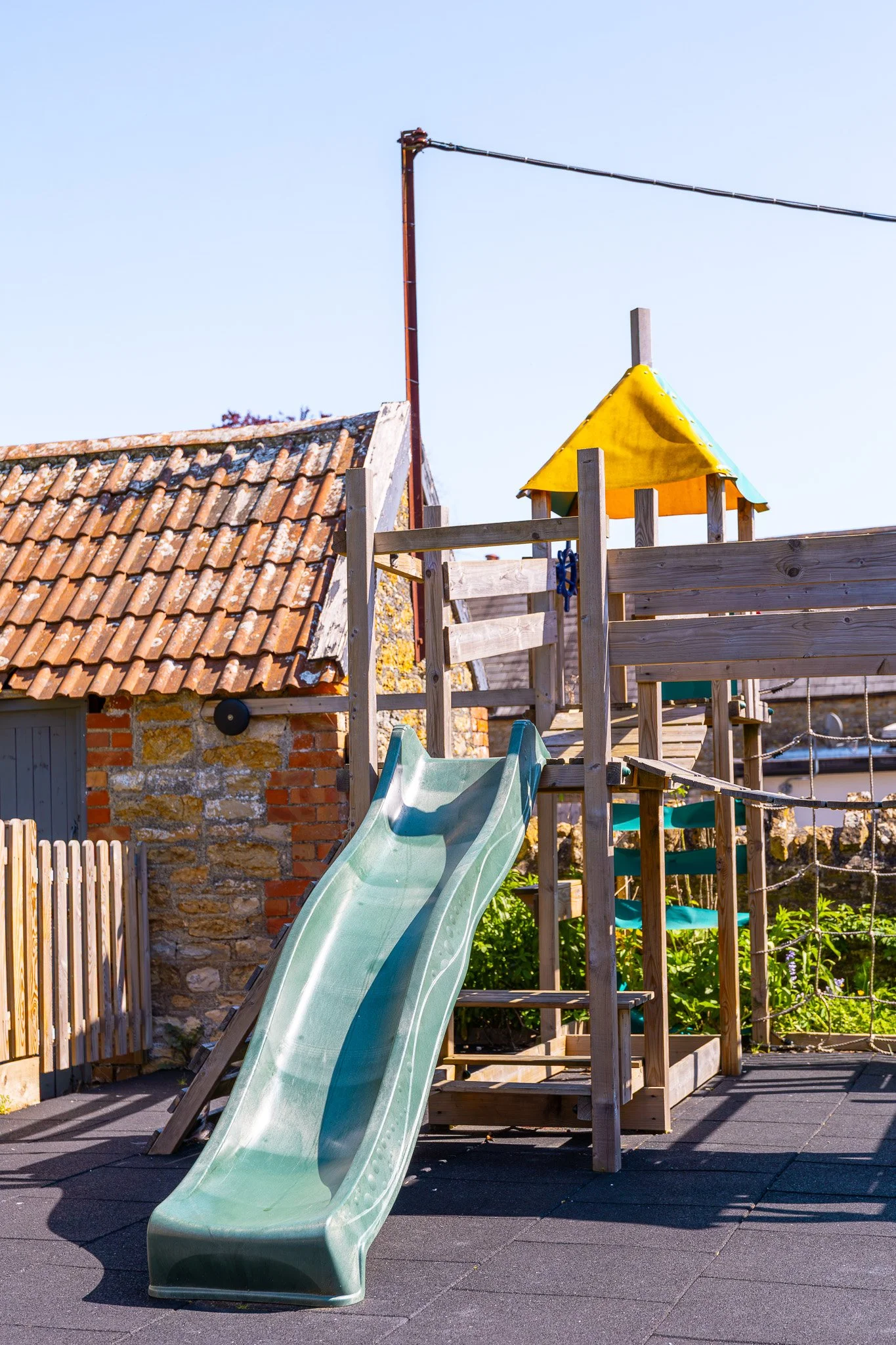 A playground with a green slide attached to a wooden structure. The background shows a building with a tiled roof and a metal pole with a wire. There's also a yellow and blue canopy on top of the wooden structure, and the ground is covered with rubbe