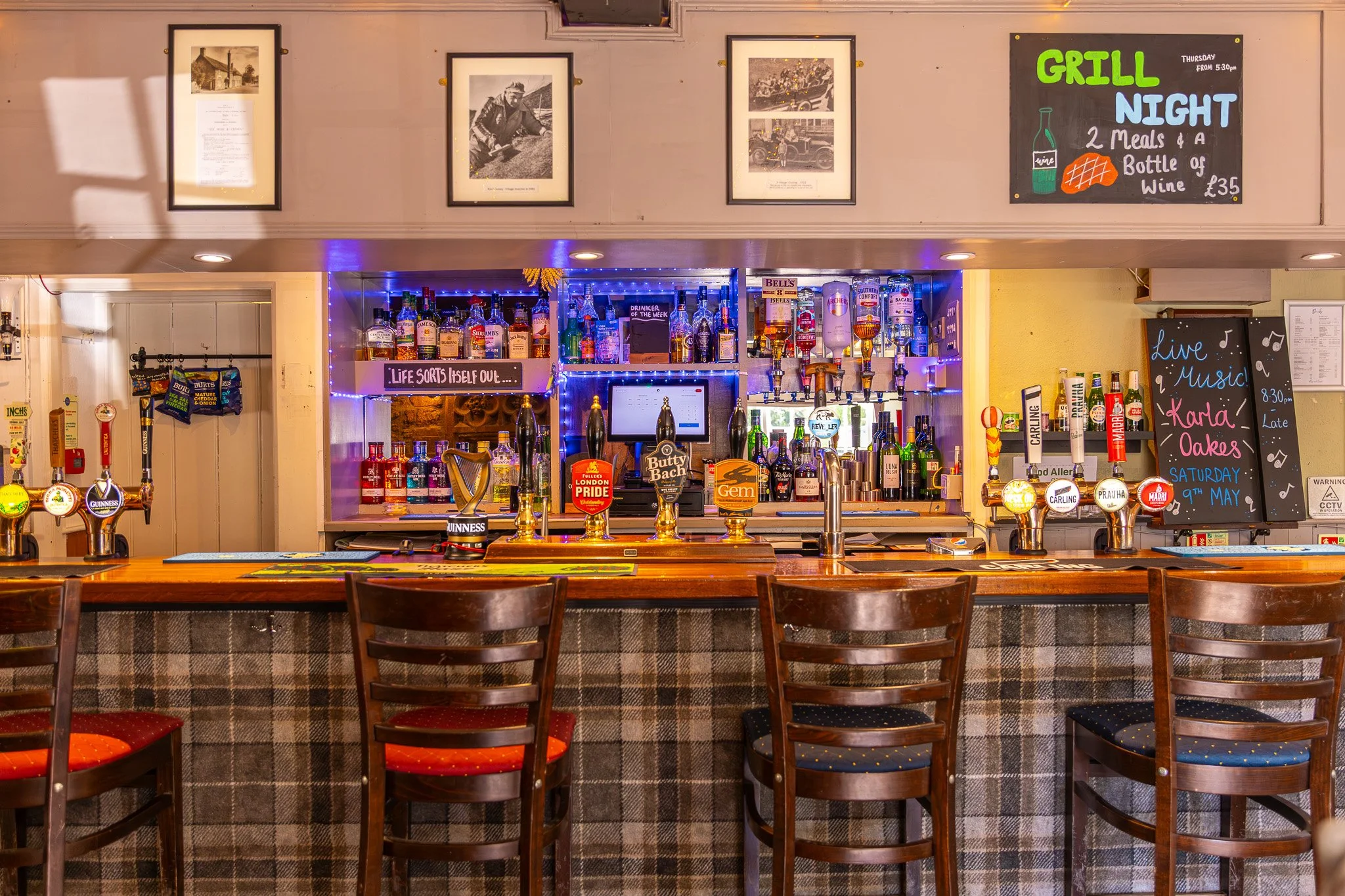 Bar interior with a wooden counter, beer taps, bottles of alcohol, and chalkboard signs advertising drinks and live music. Walls decorated with framed pictures and a chalkboard sign for a Thursday grill night and Saturday live music event.