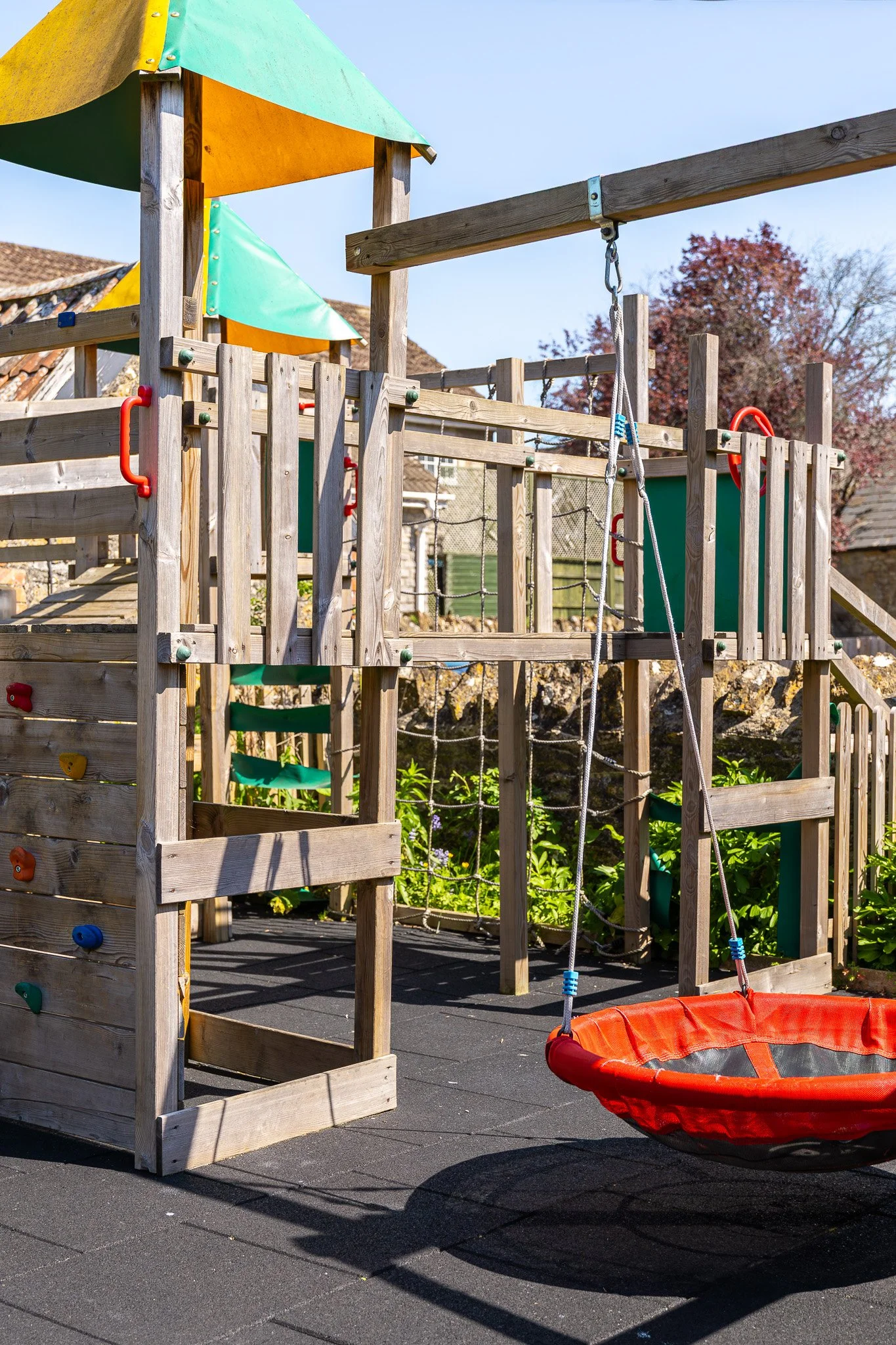 Child's wooden playground structure with green and yellow shaded roof, climbing wall, rope ladder, and red bucket swing on a black rubber surface outdoors, with trees and buildings in the background.