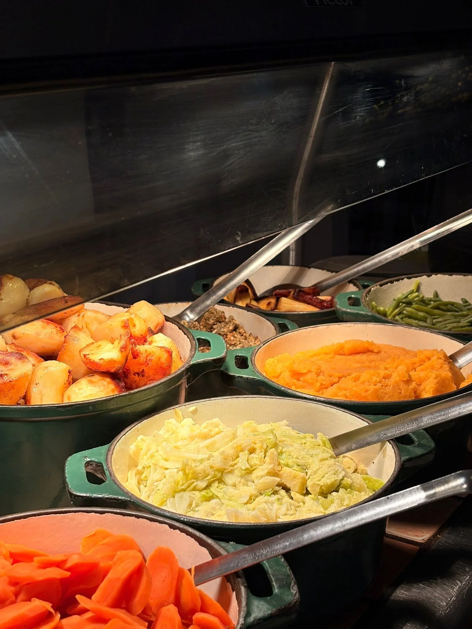 A buffet of cooked vegetables including carrots, mashed sweet potatoes, green beans, broccoli, mashed potatoes, and lettuce, with tongs for serving.