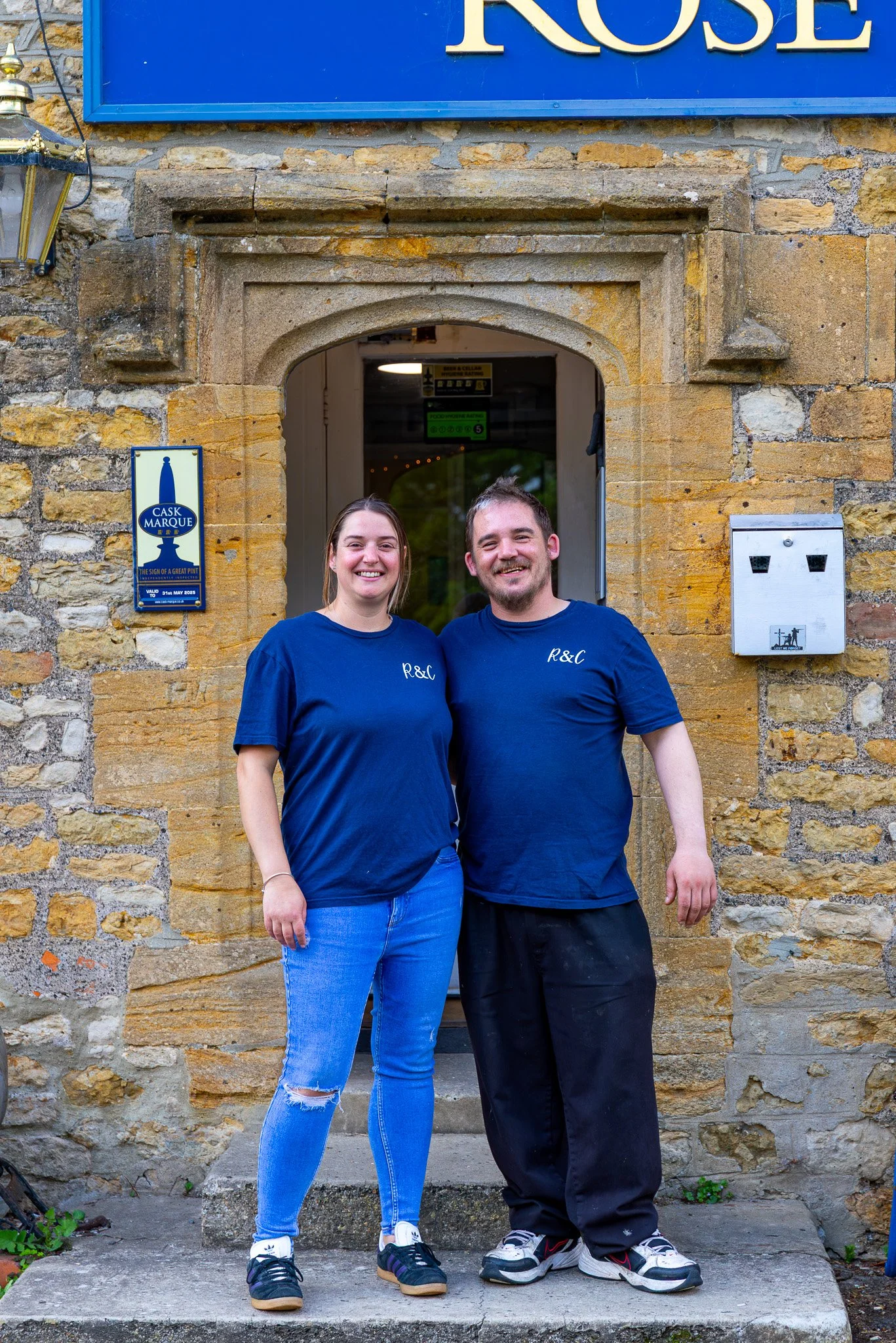 Two smiling young adults, a woman and a man, standing on a stone step in front of an old stone building with a blue sign that says 'ROSE'. They are wearing matching blue T-shirts with initials 'R&L' and casual jeans and sneakers.
