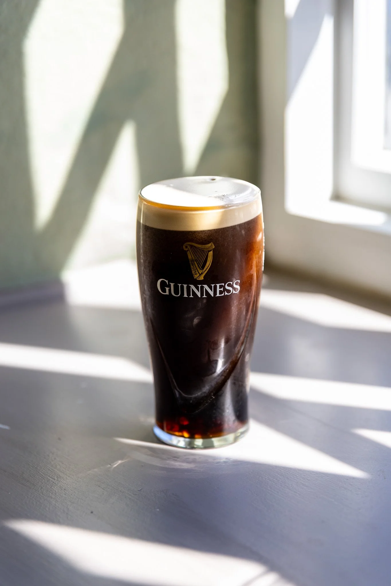 A pint glass of Guinness beer on a white surface near a window, with sunlight casting shadows on the wall behind it.