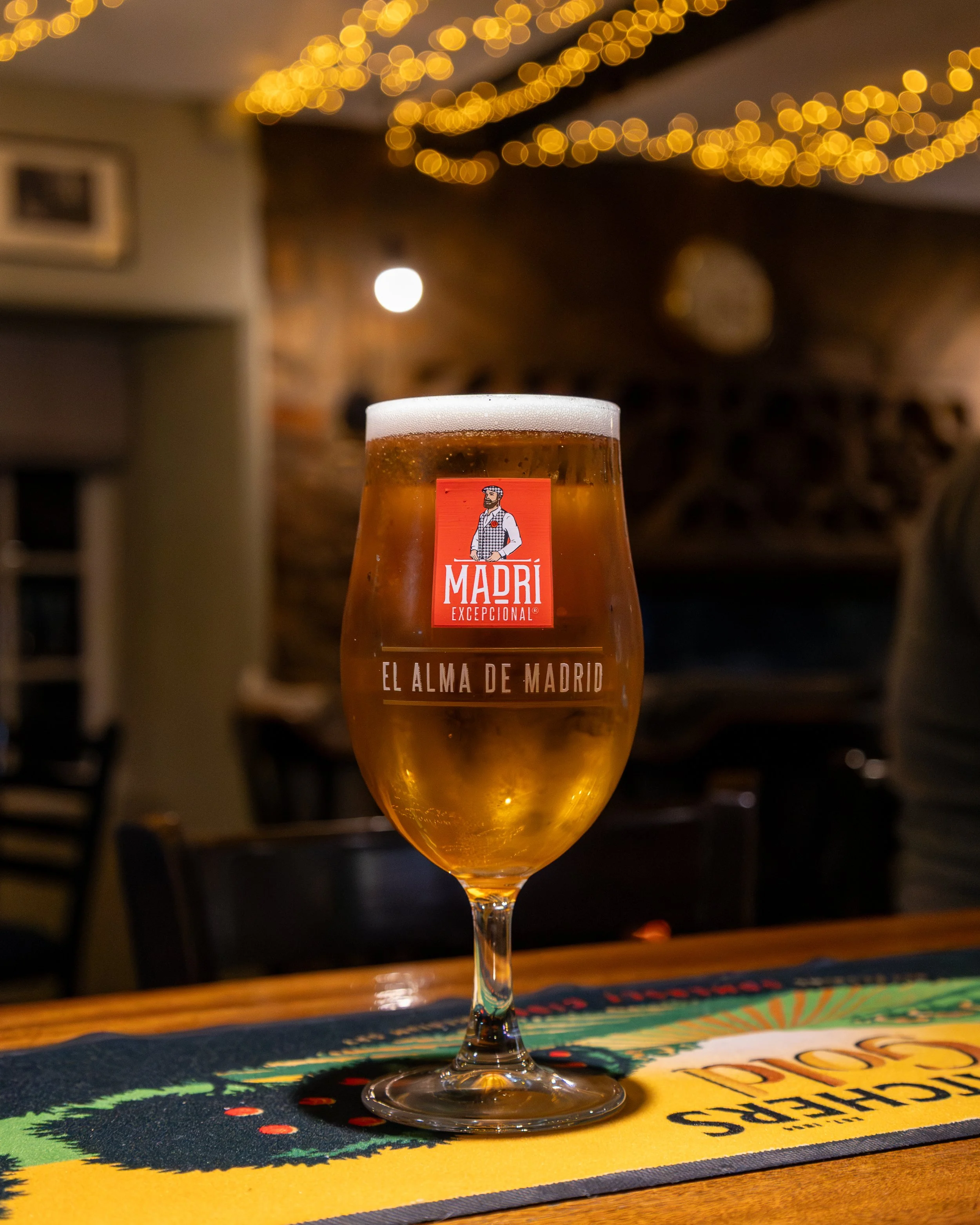 A glass of beer with a Frothy head on a colorful table mat in a dimly lit bar or restaurant with string lights hanging from the ceiling.