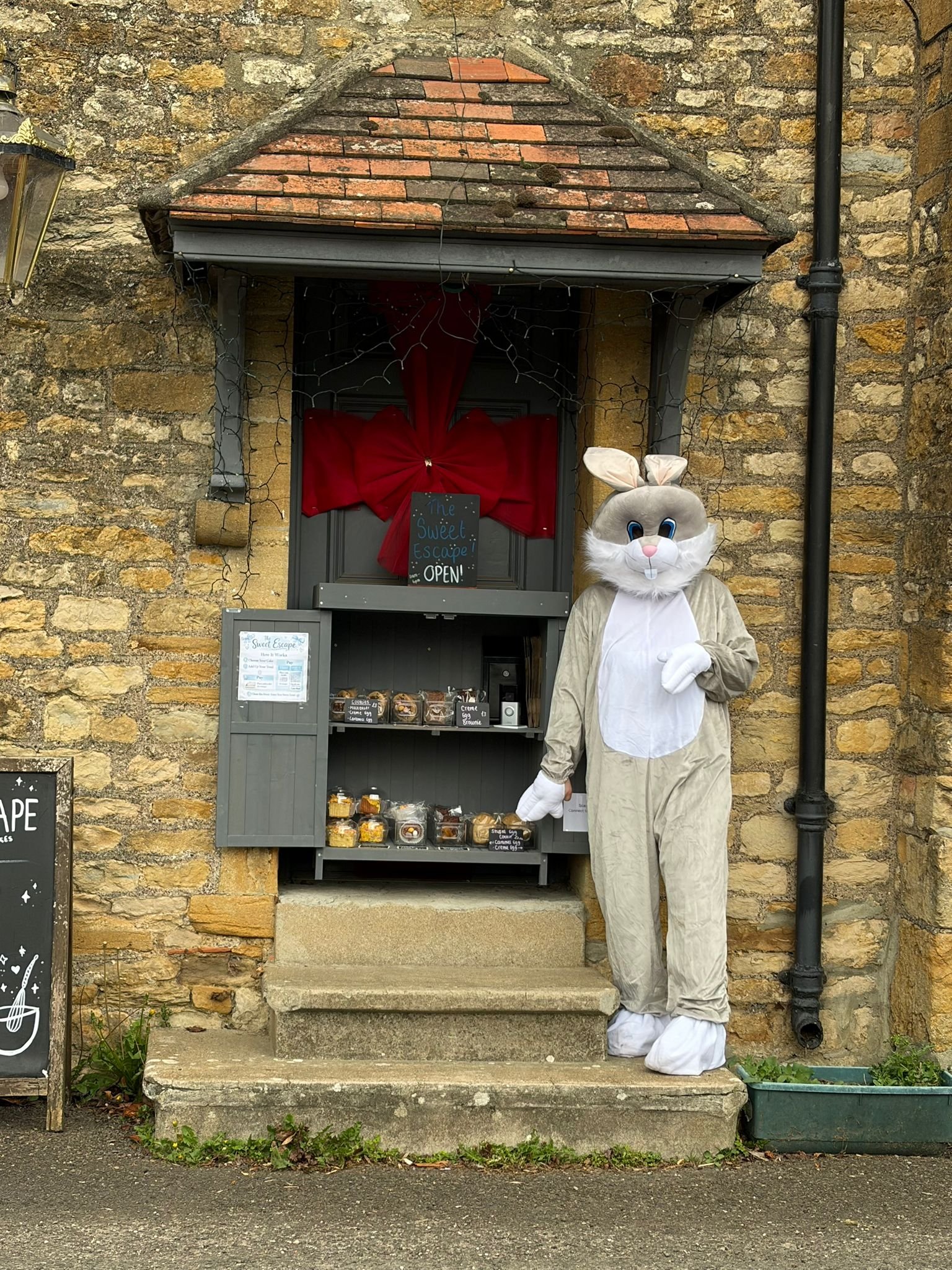 A person dressed in a bunny costume standing in front of a small kiosk on a stone doorstep. The kiosk has baked goods on display and a sign that reads 'The Sweet Escape OPEN!' The background features a brick building with a small roof and a black dow